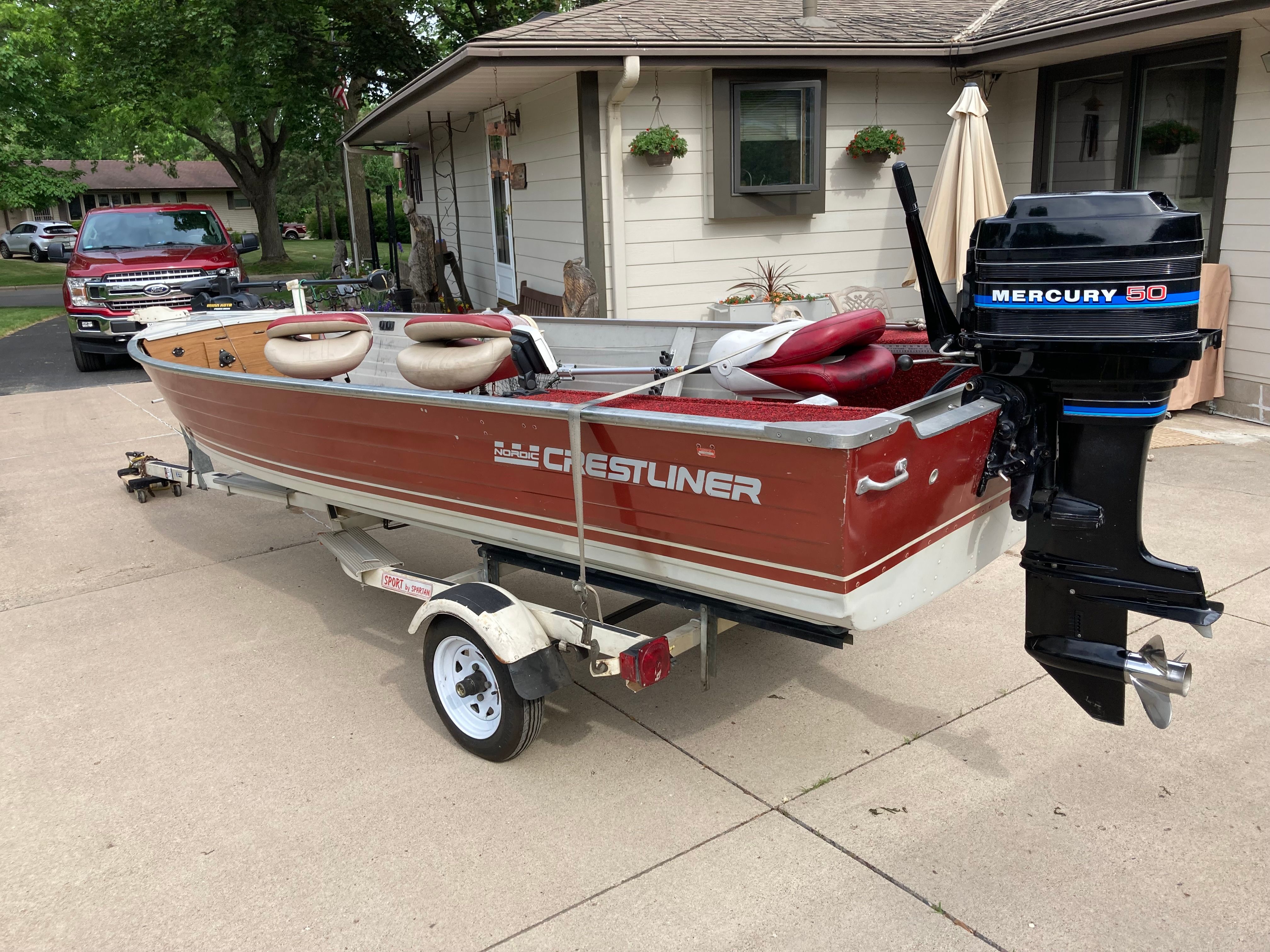 A red Crestliner boat with a Mercury motor is on a trailer in a driveway.