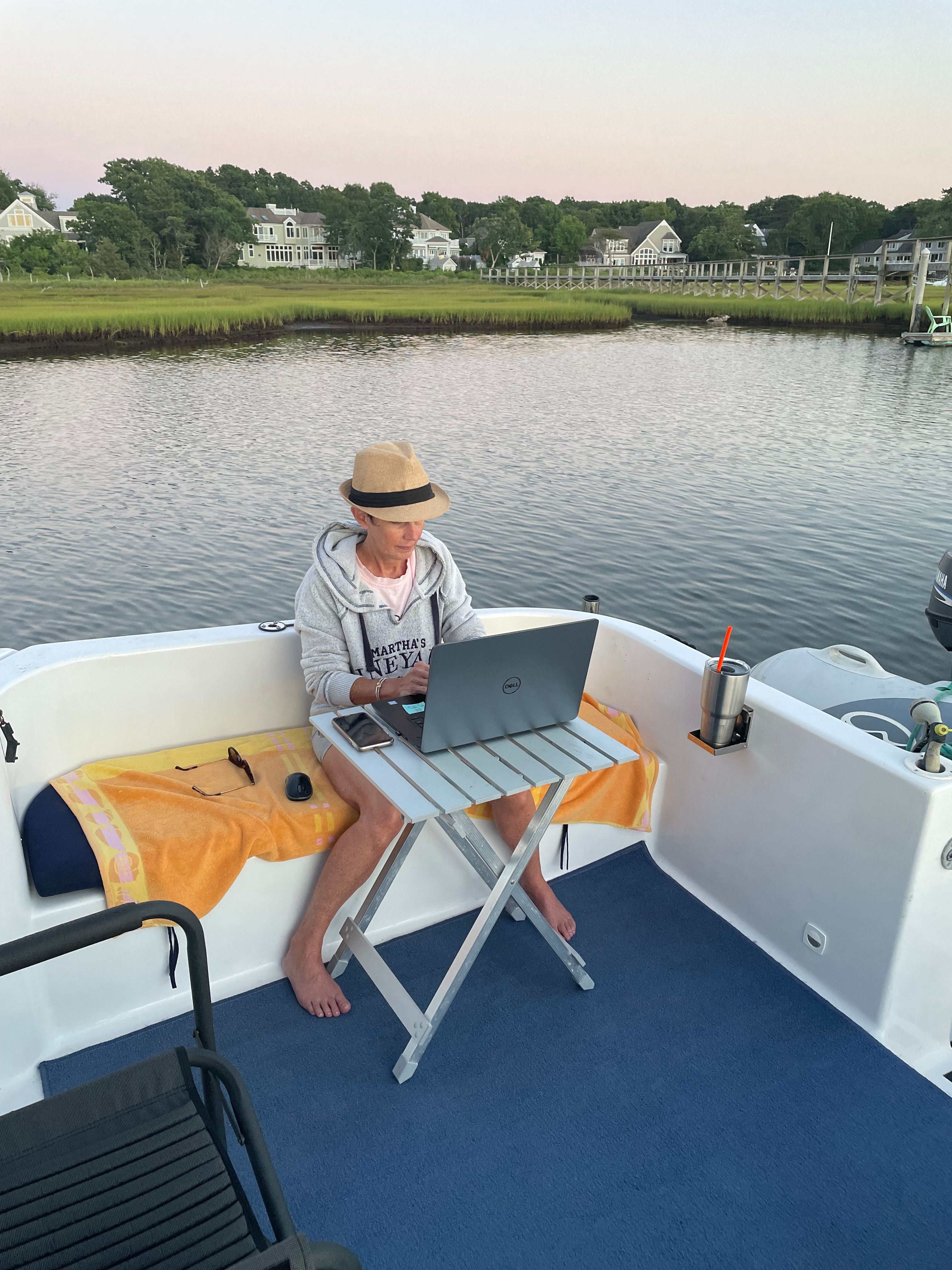 A person works on a laptop on a boat overlooking calm water and houses.