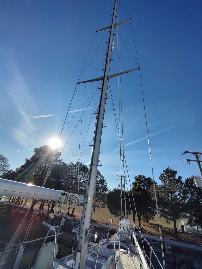 Sailboat mast of 1977 Hallberg-Rassy 41 under clear blue sky with sun and trees.