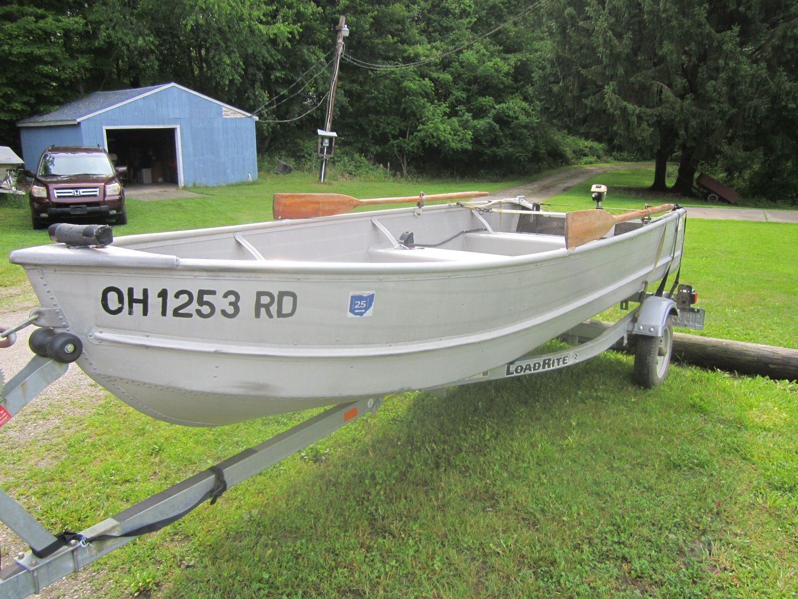 A gray boat on a trailer sits on grass in front of a blue shed.