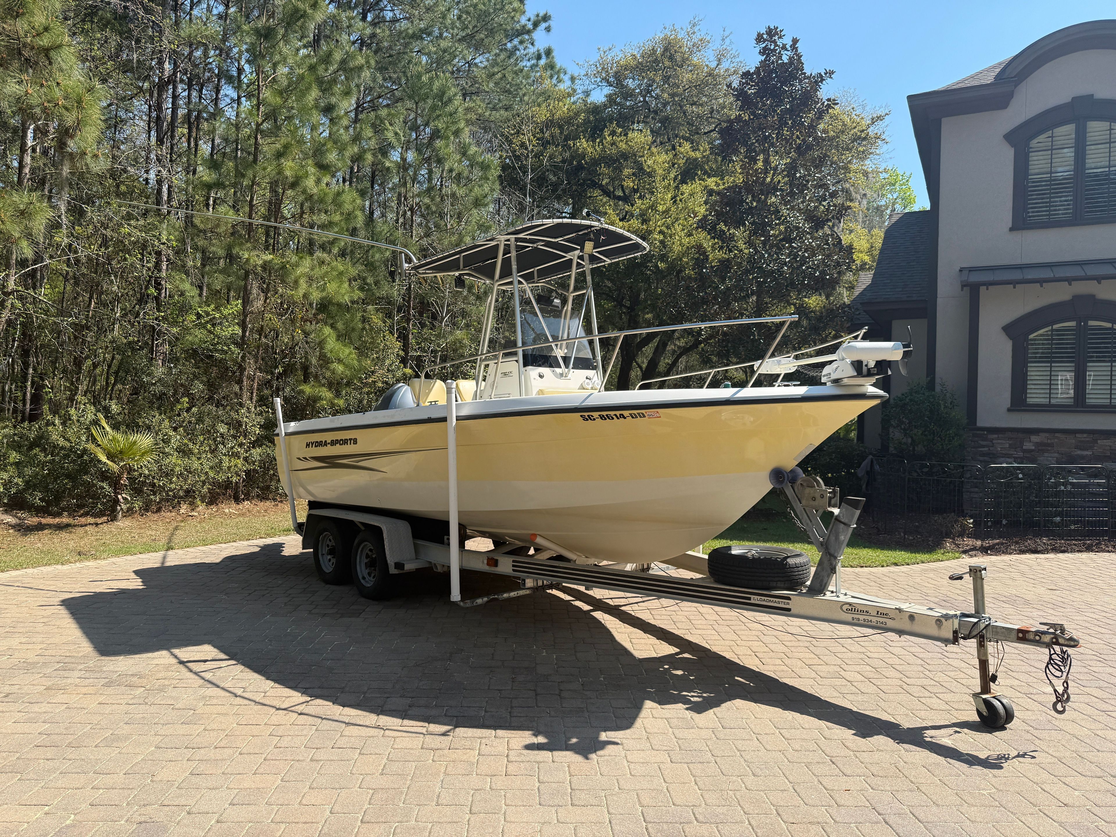 A yellow boat on a trailer parked in front of a house and trees.