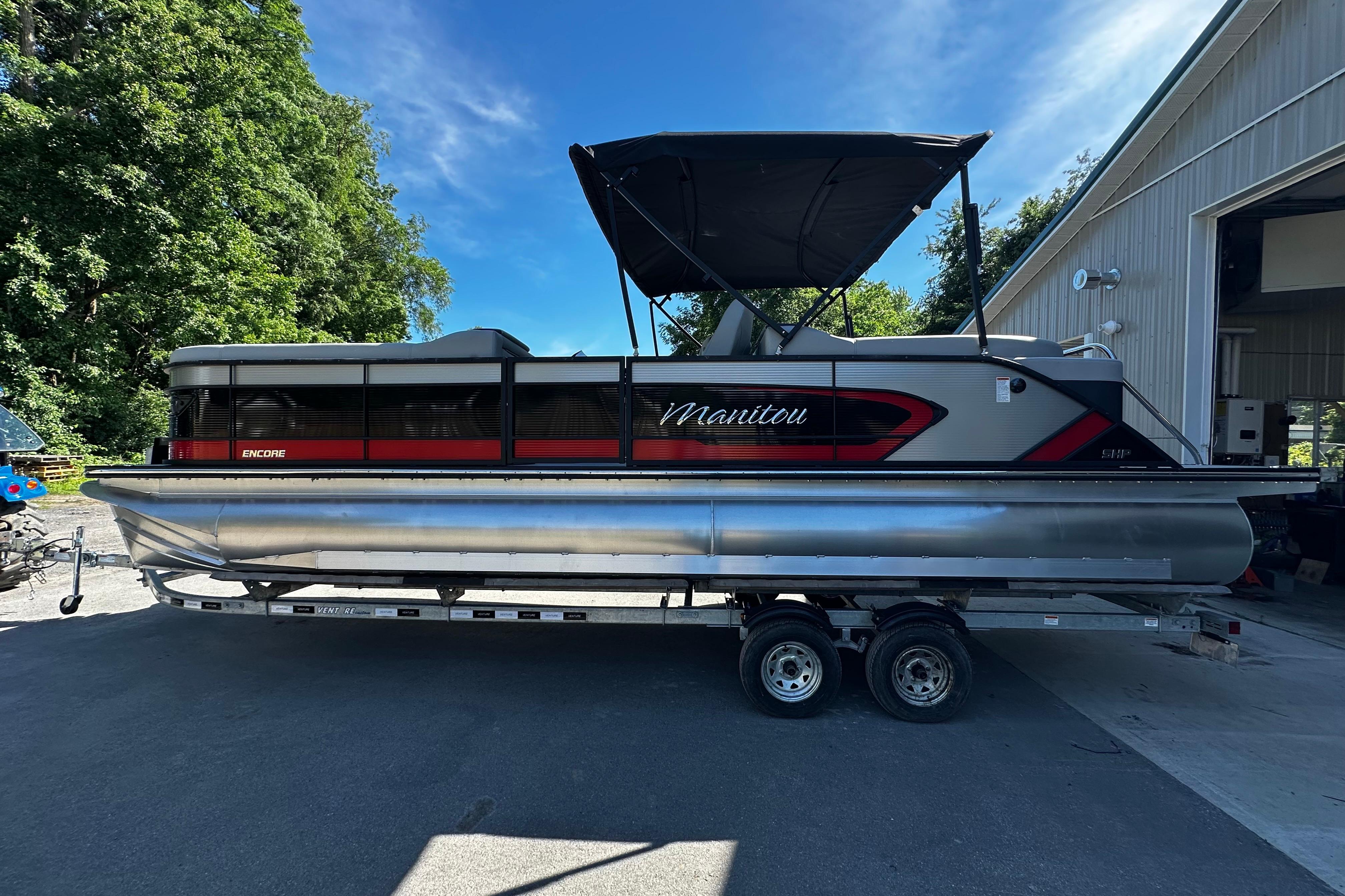 2027 Manitou 25 Encore RF pontoon boat on trailer, parked near a building.