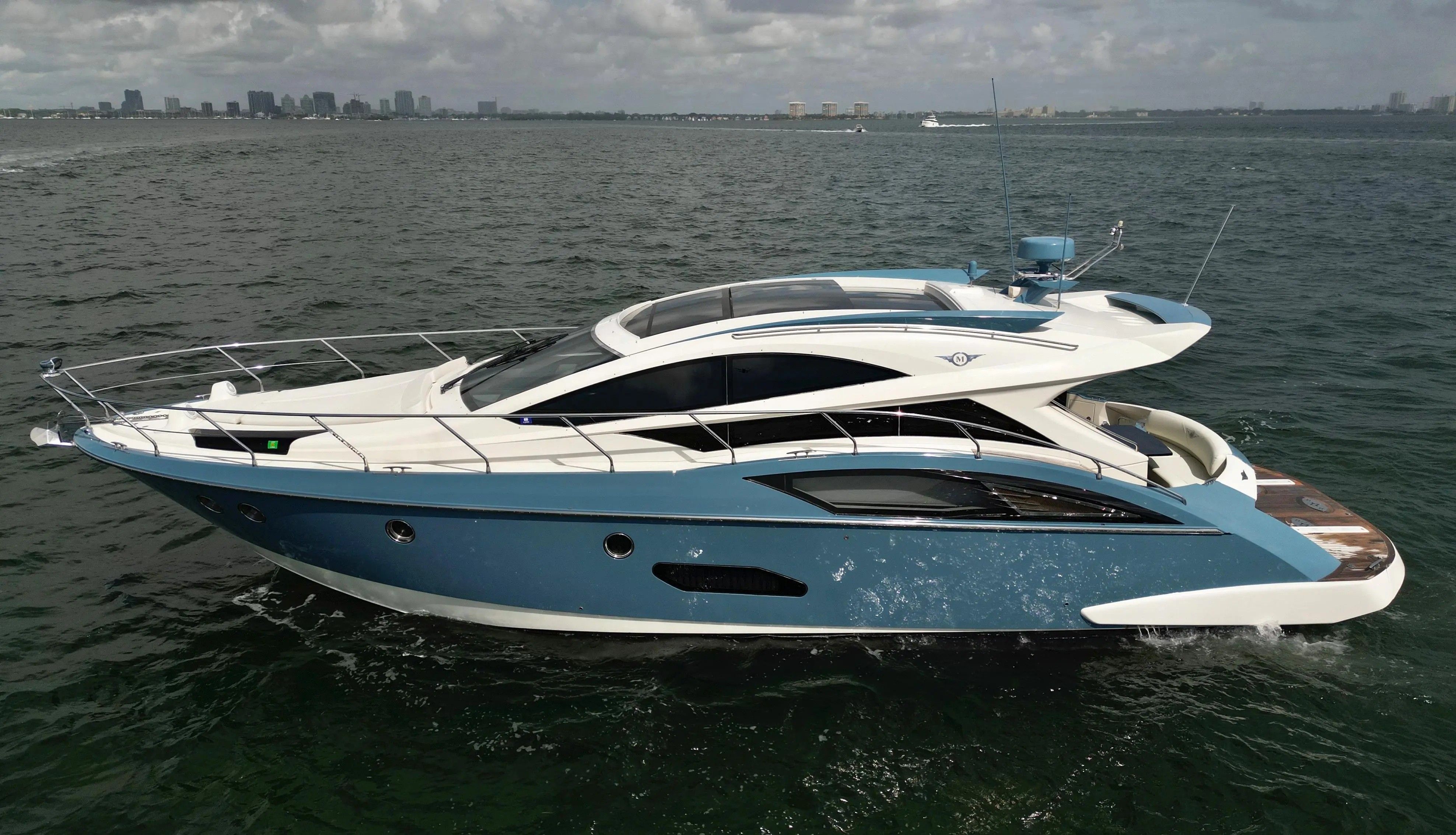 A sleek blue and white yacht cruises on the sea with a city skyline in the background.