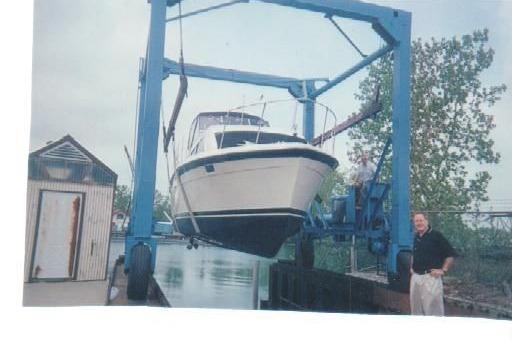 1977 Trojan 36 Tri-Cabin boat lifted by crane near water, with a person standing nearby.