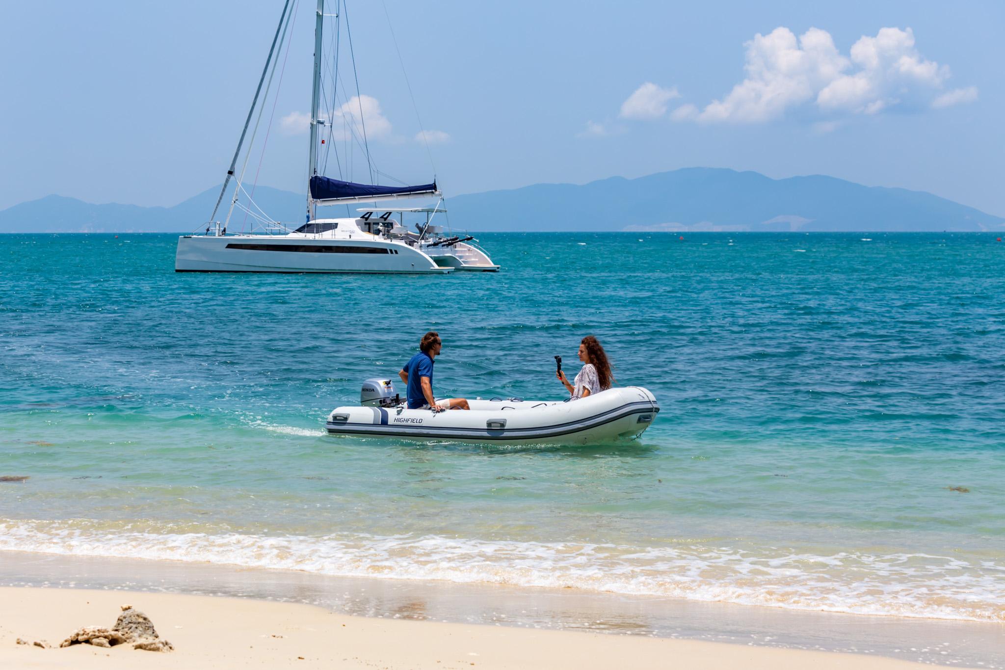 Couple in dinghy near Seawind 1600 Passagemaker catamaran, 2025, anchored by tropical beach.