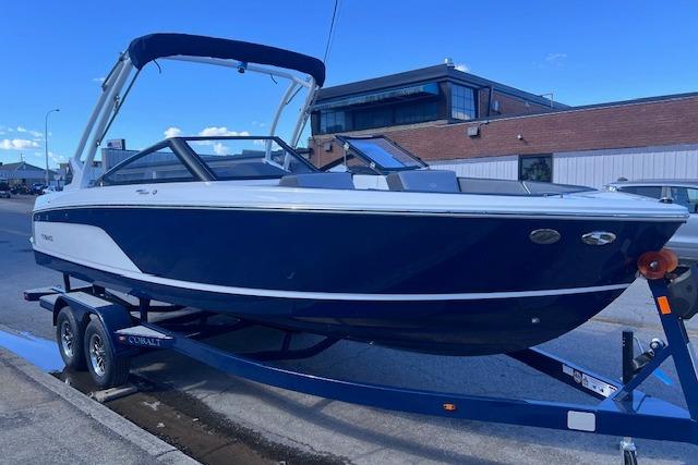 2025 Cobalt CS23 Surf boat on trailer, parked outdoors, blue and white design.