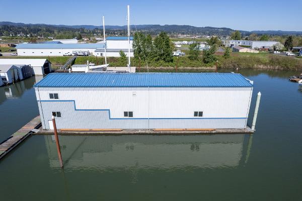 Aerial view of 1985 Custom Larson Boathouse on calm water, blue roof, industrial background.