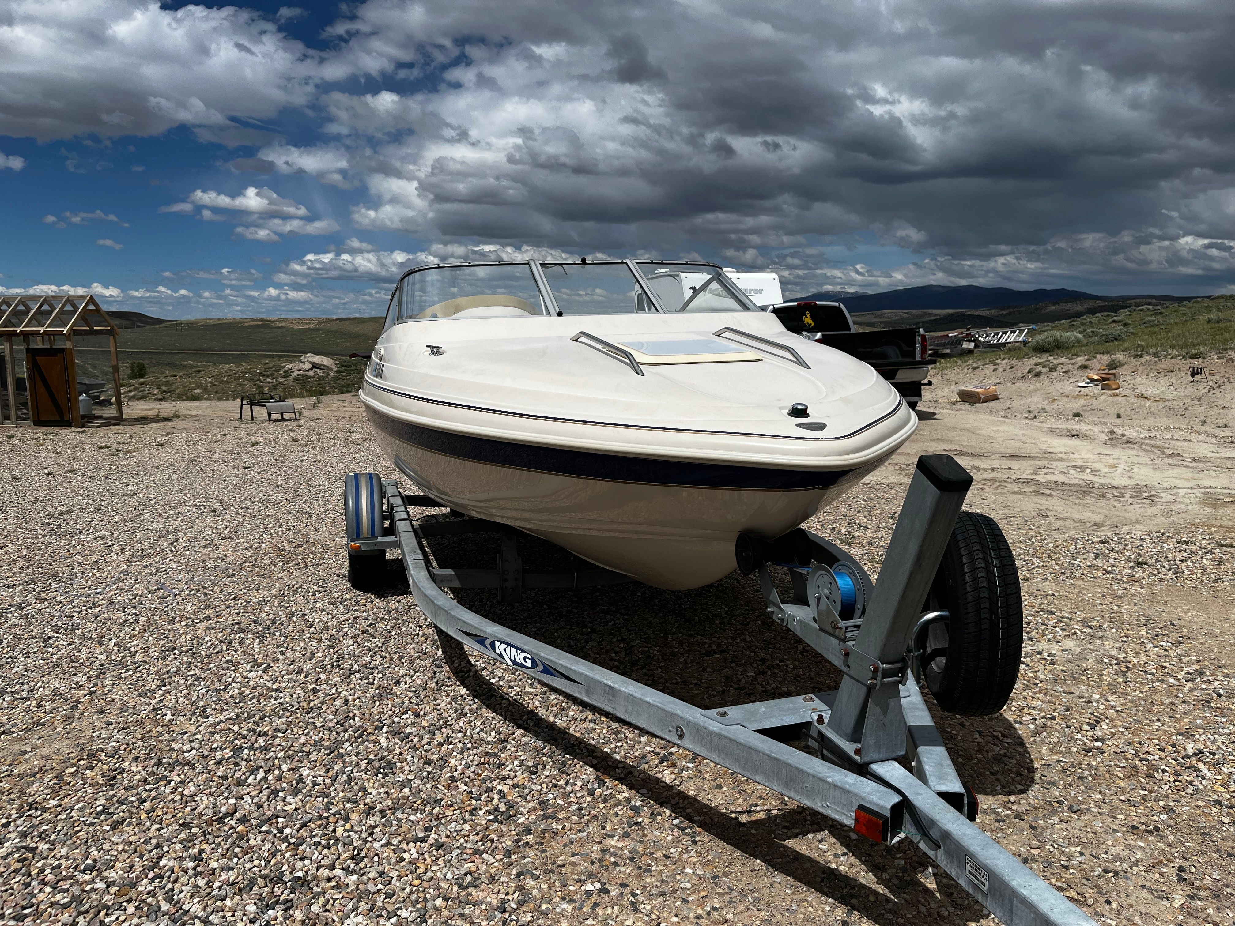 A 1998 Glastron SE199 boat on a trailer on a gravel lot under a cloudy sky.