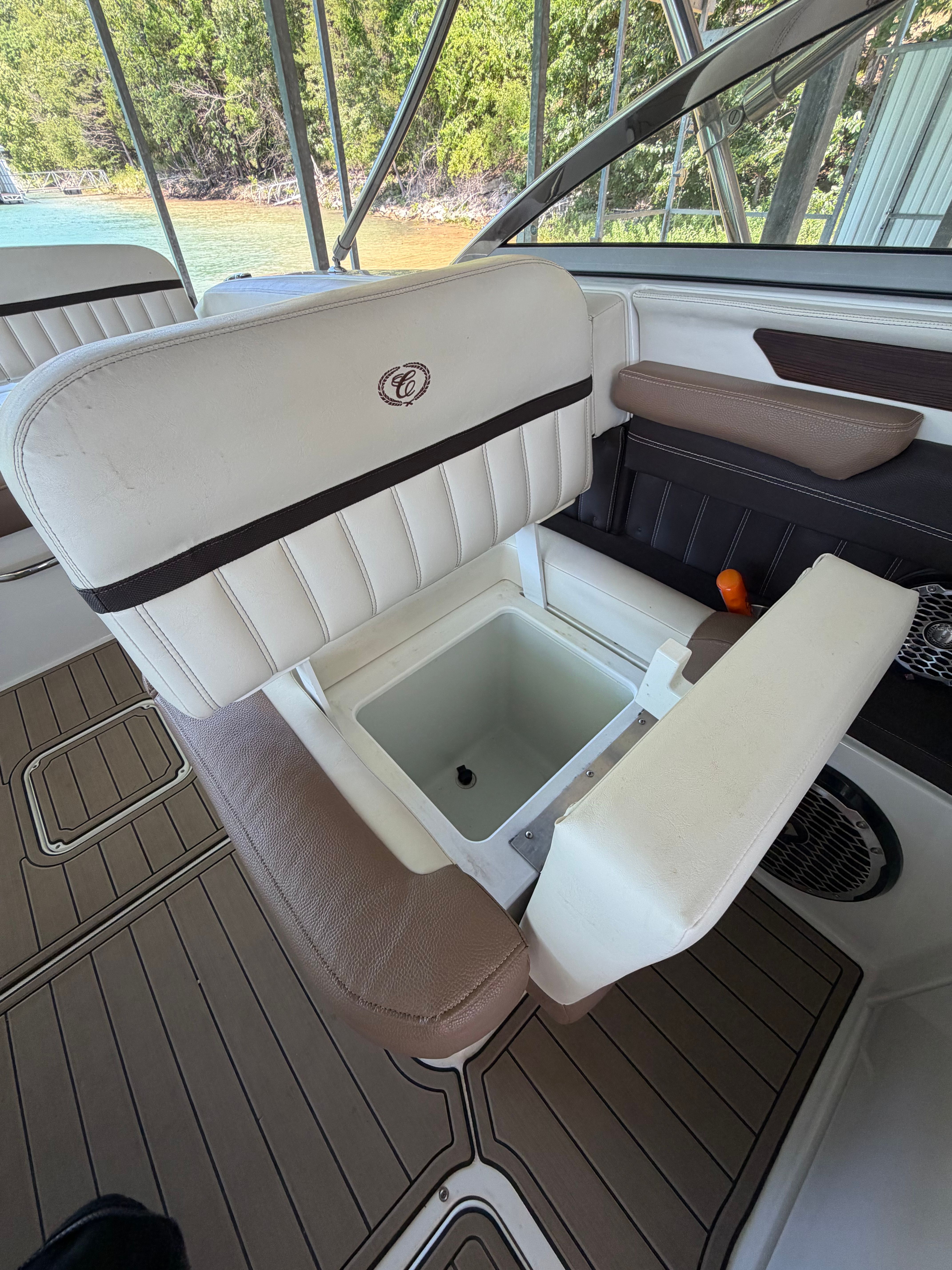 An open seat with storage on a boat, surrounded by trees and water.