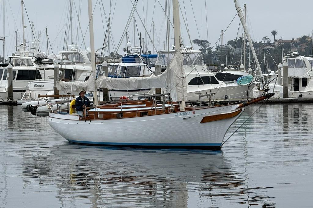 Vintage 1965 sailboat in marina, surrounded by yachts and calm water reflections.