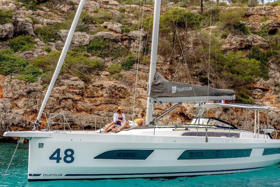 2026 Dufour 48 yacht anchored near rocky coastline with people relaxing on deck.
