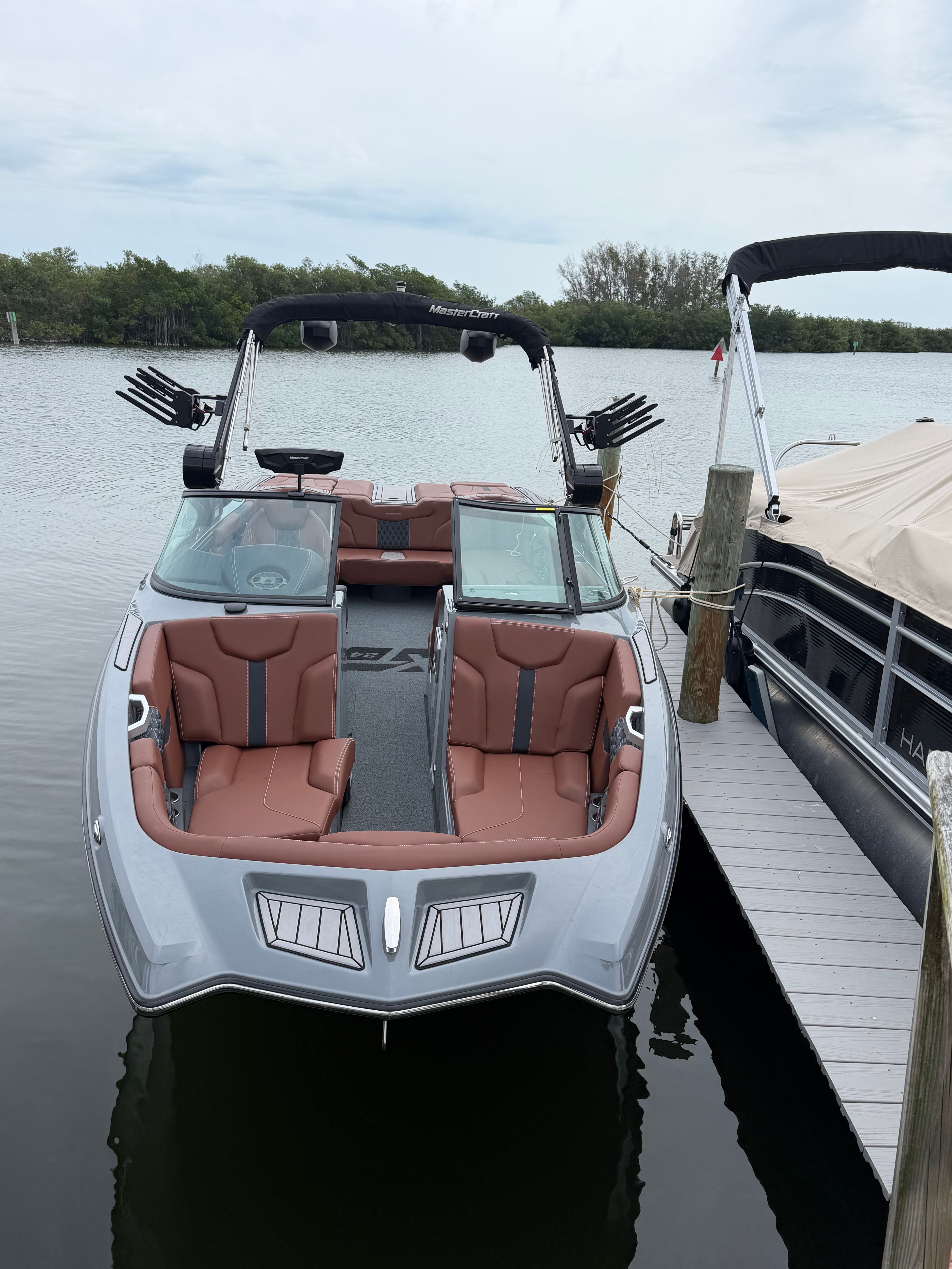 A MasterCraft XT24 boat docked at a pier, surrounded by calm water and trees.