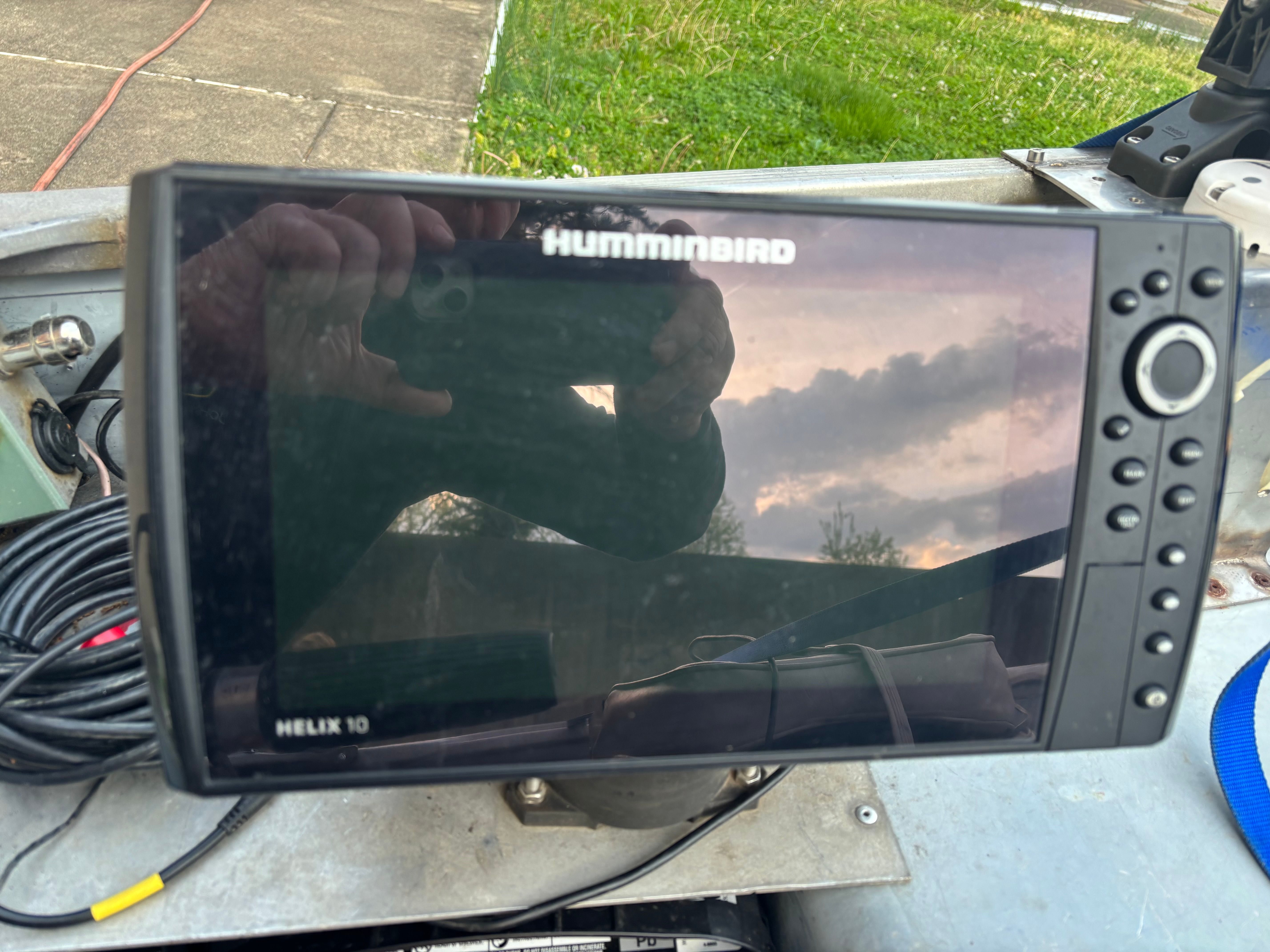 A Humminbird Helix 10 fish finder on a boat, with cables and a cloudy sky reflected.