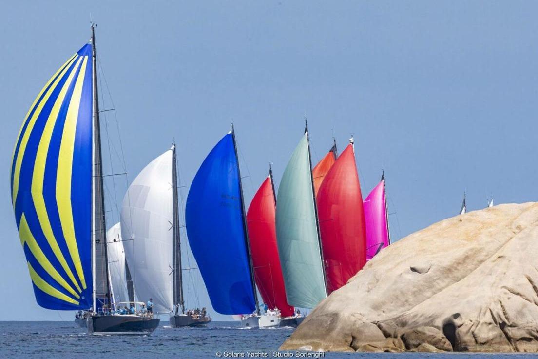 Colorful sailboats, including Solaris 55 (2022), racing near rocky coastline.
