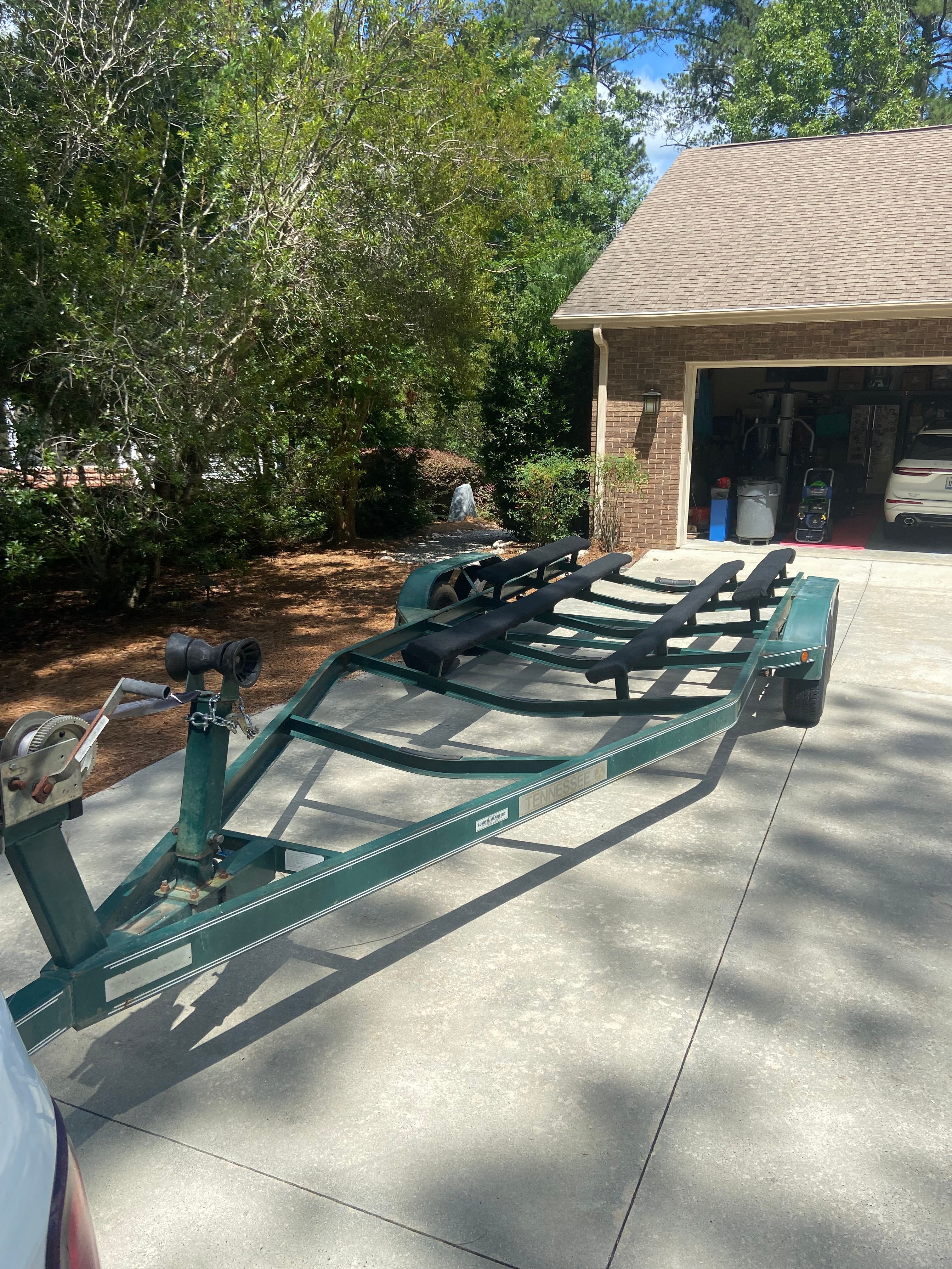 A boat trailer is parked on a driveway in front of a garage. Trees and shrubs in the background.