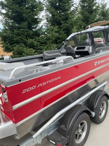 2025 Spartan 200 Astoria boat with red exterior on a trailer, surrounded by trees.