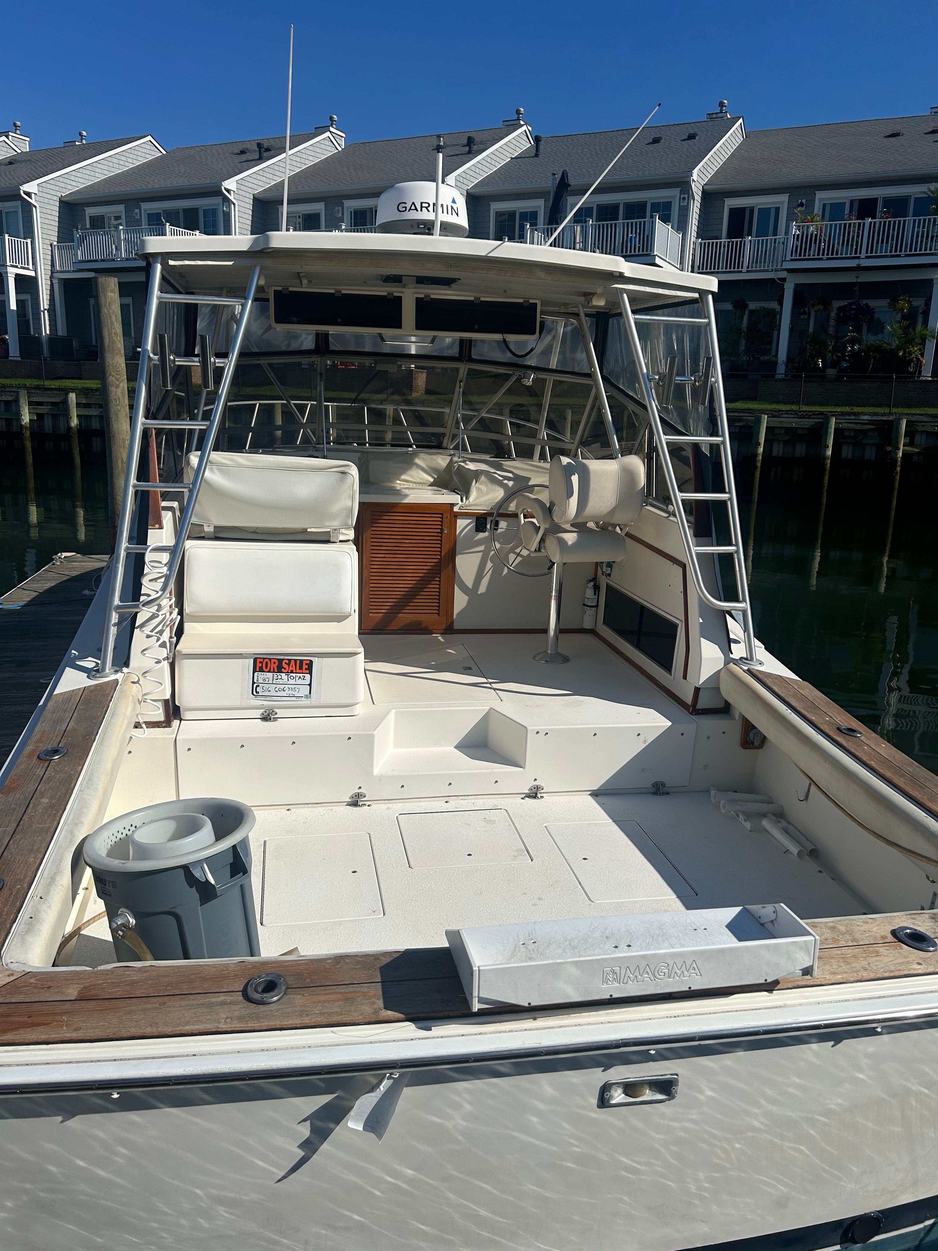 A 1987 Topaz Express boat with an open cockpit, white seating, and helm. It's docked near townhouses.