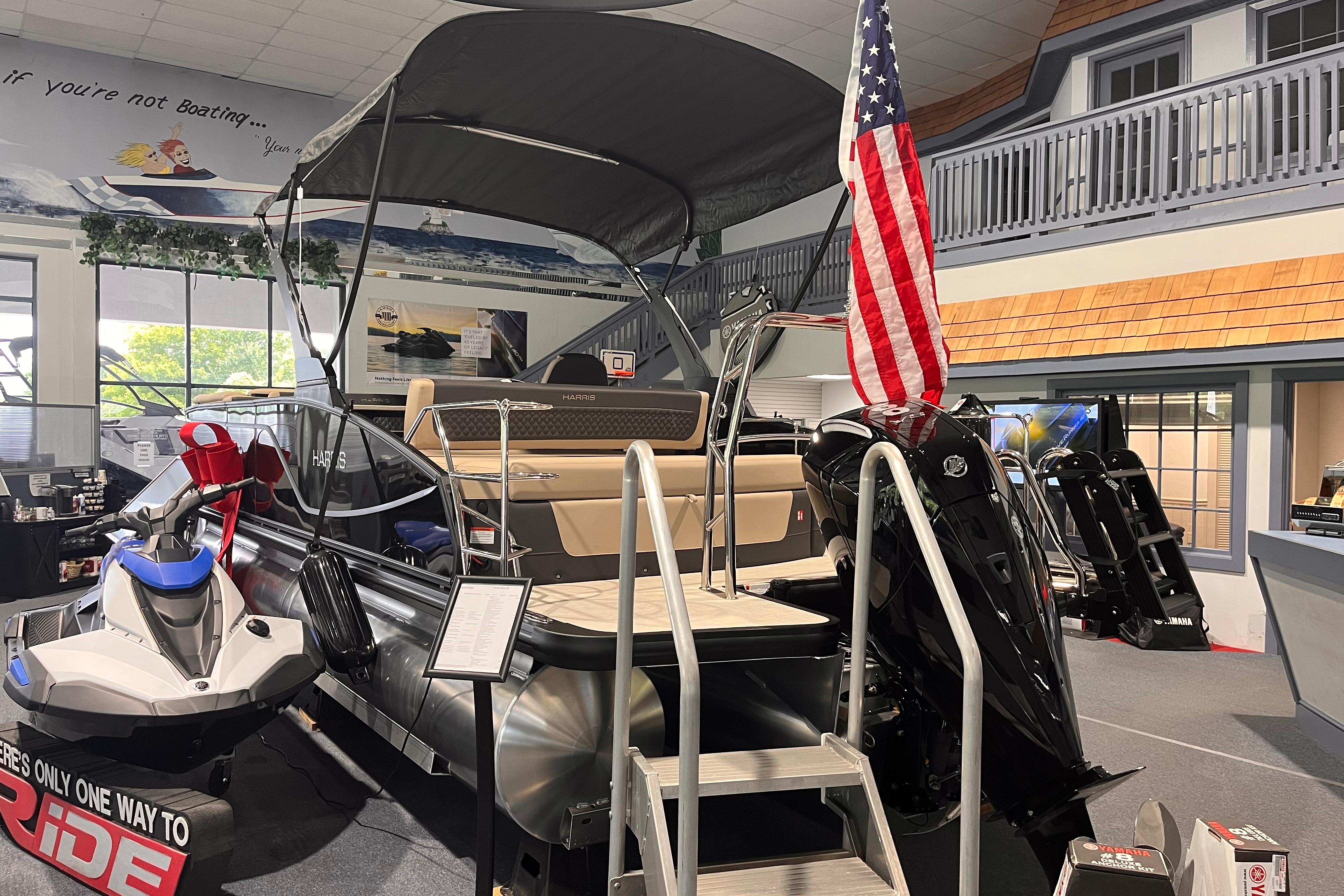 2026 Harris Sunliner 250 pontoon boat displayed indoors with American flag and jet ski.