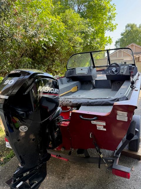 A red boat with a Mercury engine, parked on a trailer, surrounded by trees.