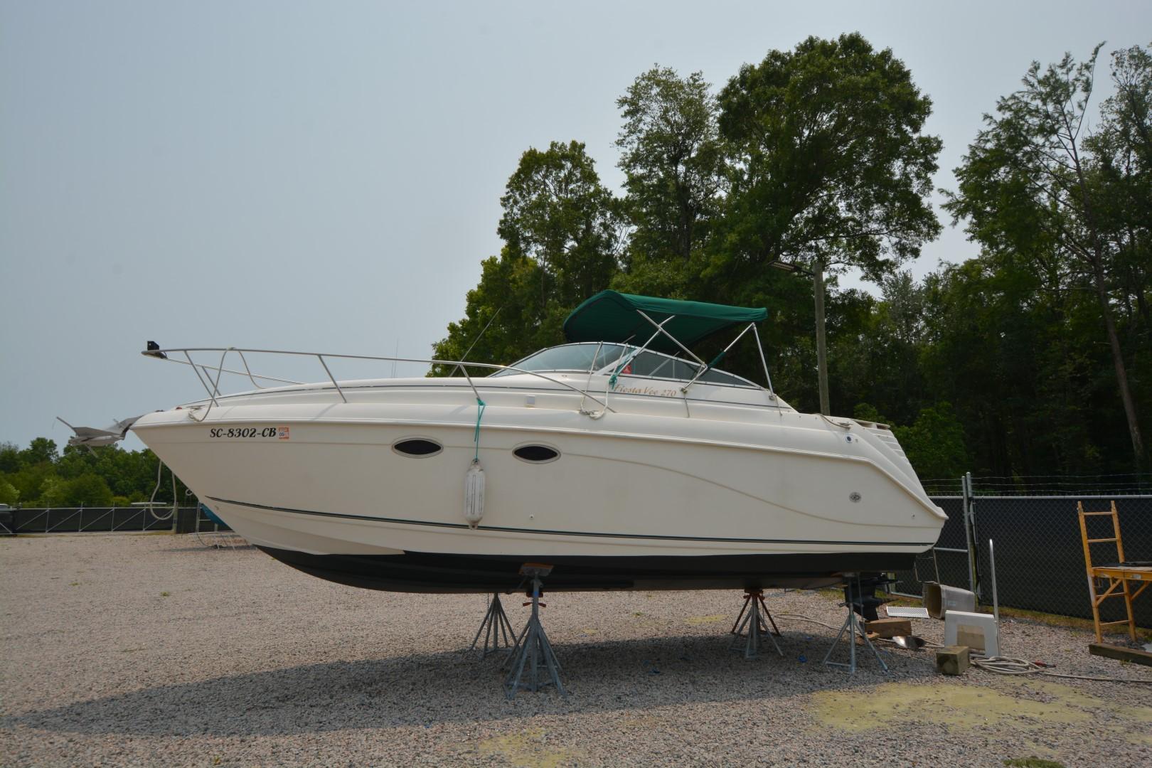 1999 Rinker 270 Fiesta Vee boat on stands, with green canopy, in a gravel lot.