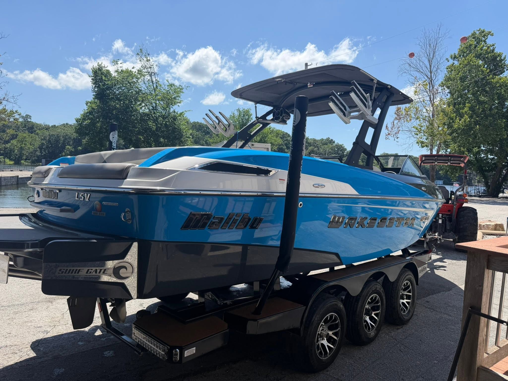 A blue Malibu Wakesetter 23 LSV boat on a trailer, surrounded by trees and a waterway.