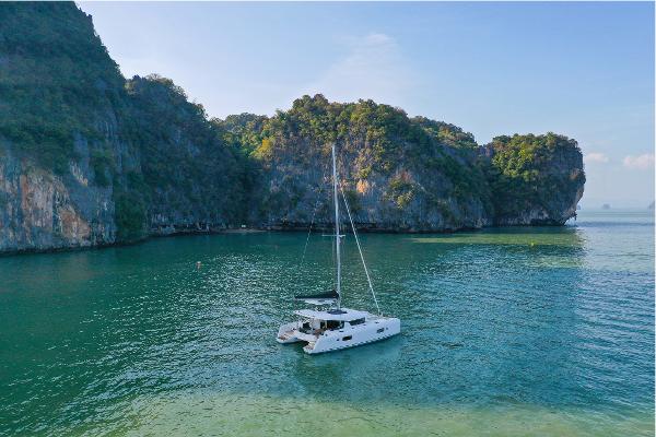 2025 Lagoon 42 Millenium catamaran sailing in a serene, tropical bay with lush cliffs.