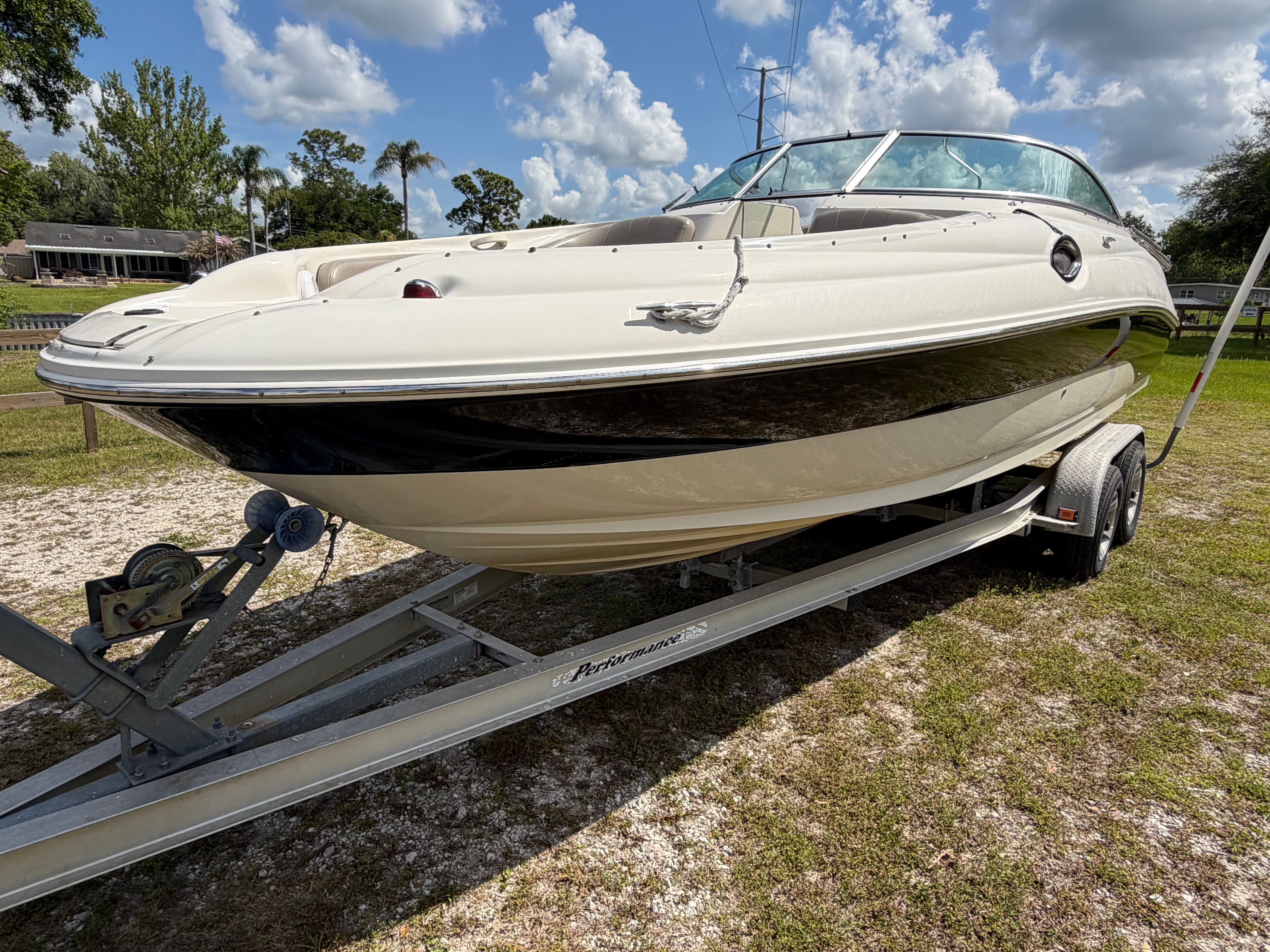 A cream-colored bowrider on a trailer, surrounded by grass and trees, under a cloudy sky.