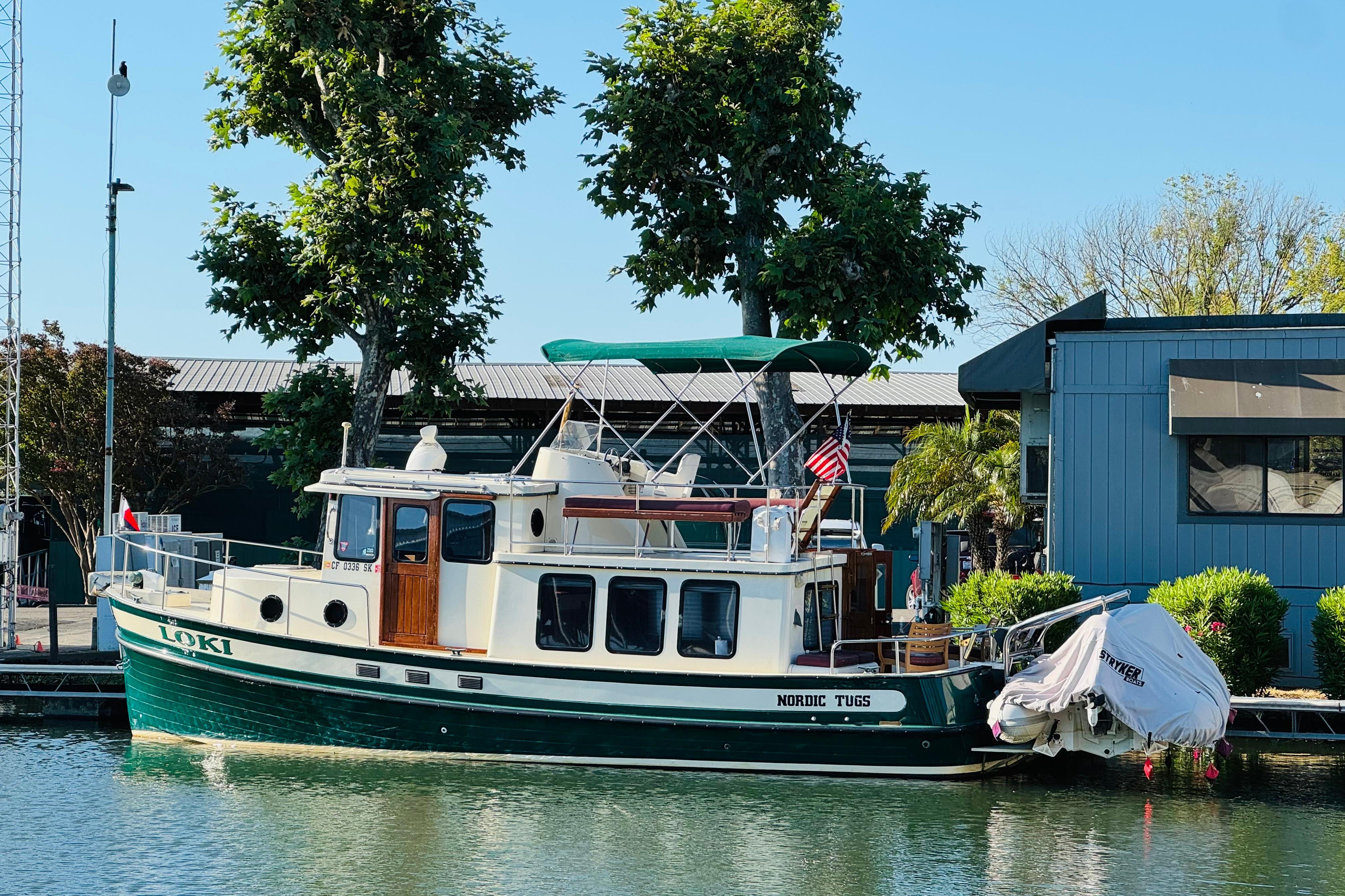 1996 Nordic Tug 32 docked by a modern building, surrounded by trees and calm water.