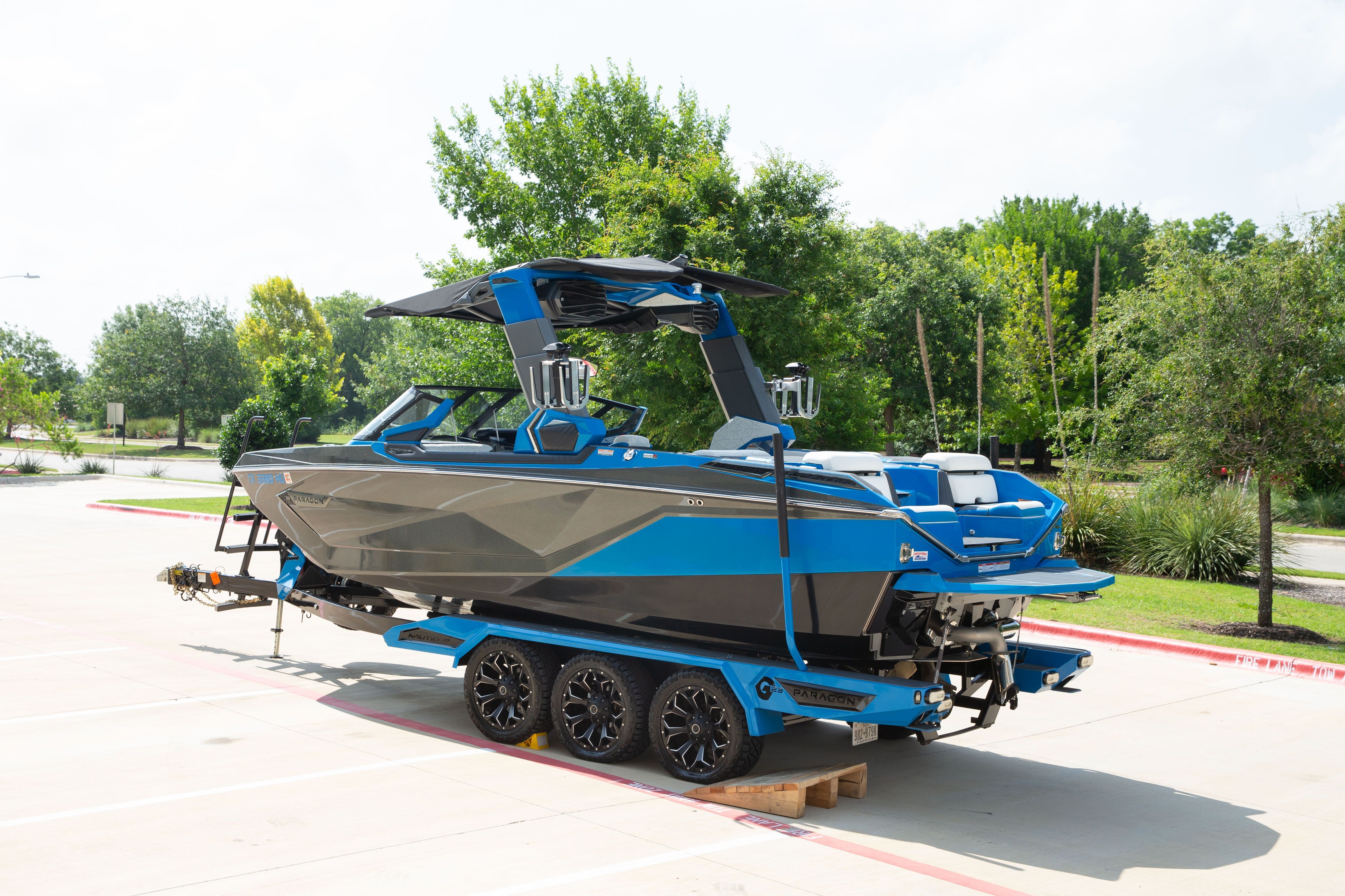 A blue boat on a trailer in a parking lot, surrounded by trees.