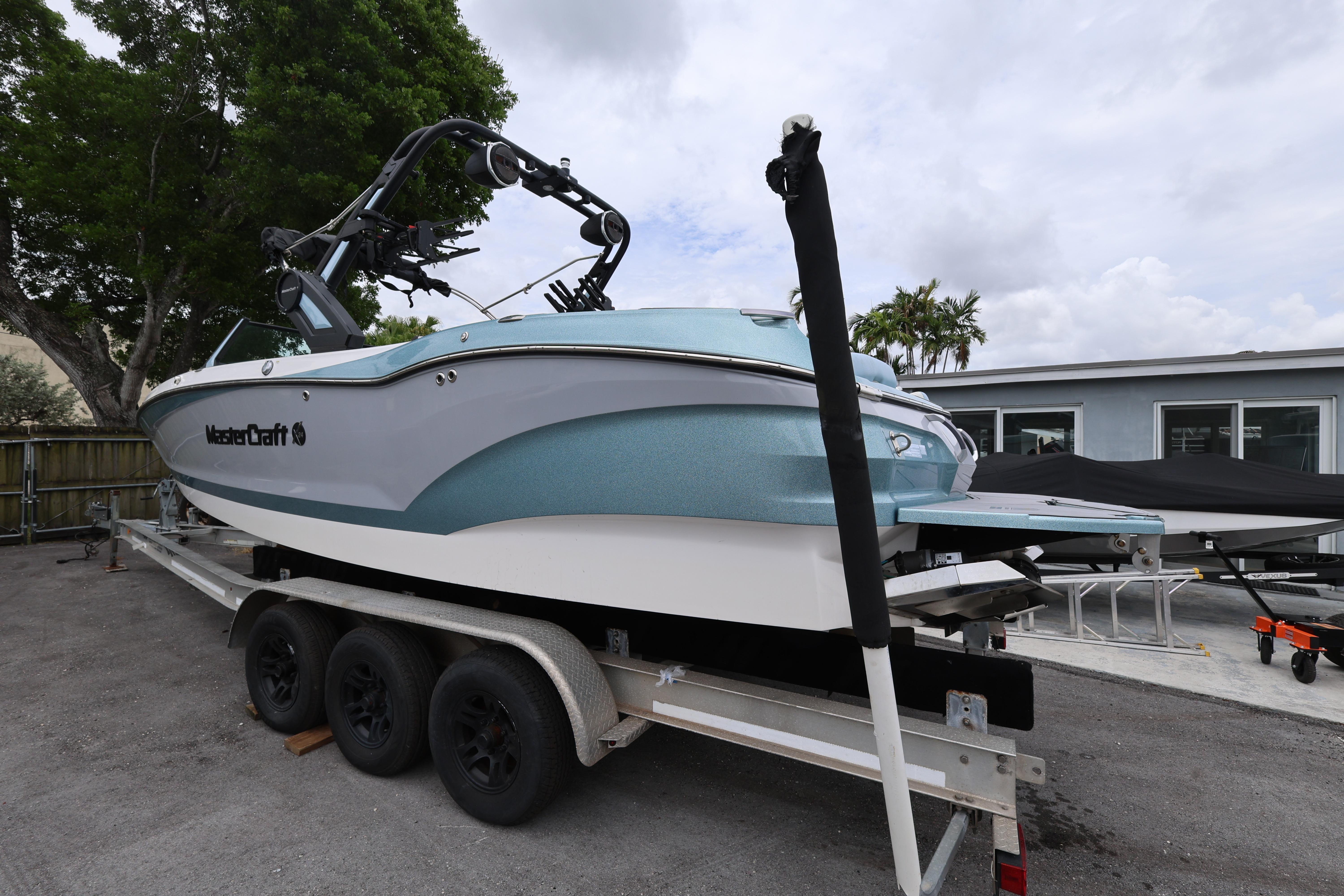 2023 MasterCraft X24 boat on trailer, parked outdoors near trees and buildings.
