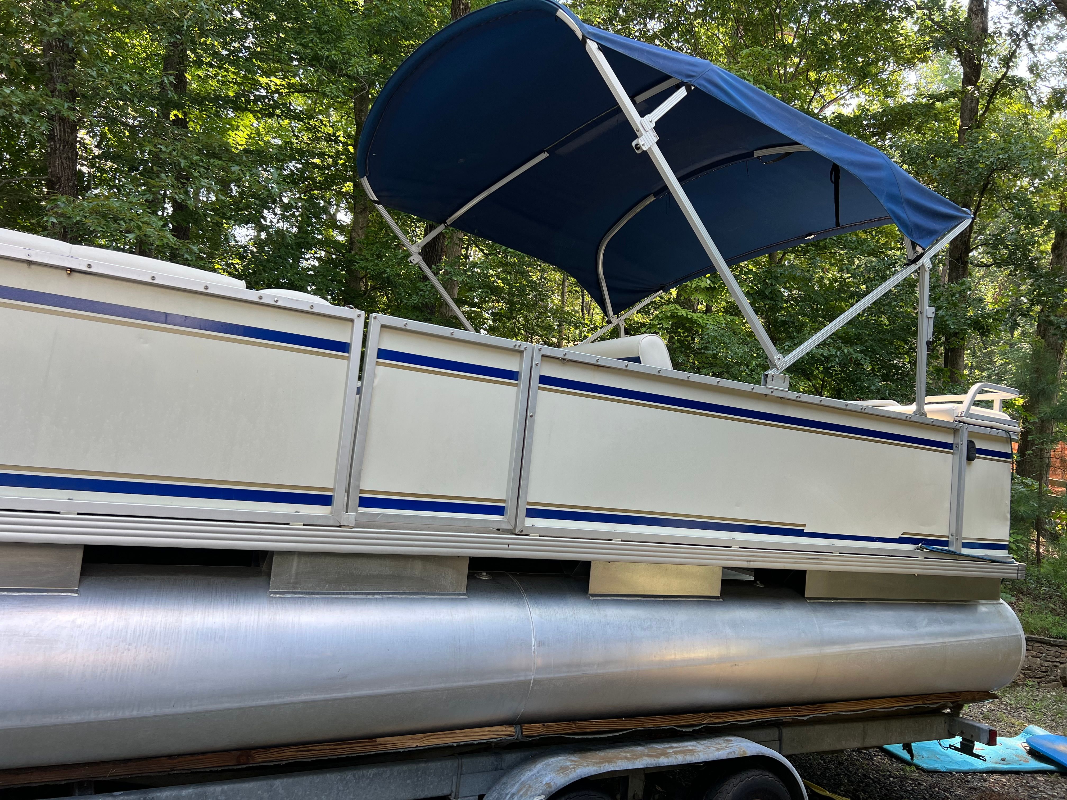 A 2003 Crest 24’ Sport pontoon boat with a blue canopy, on a trailer in the woods.