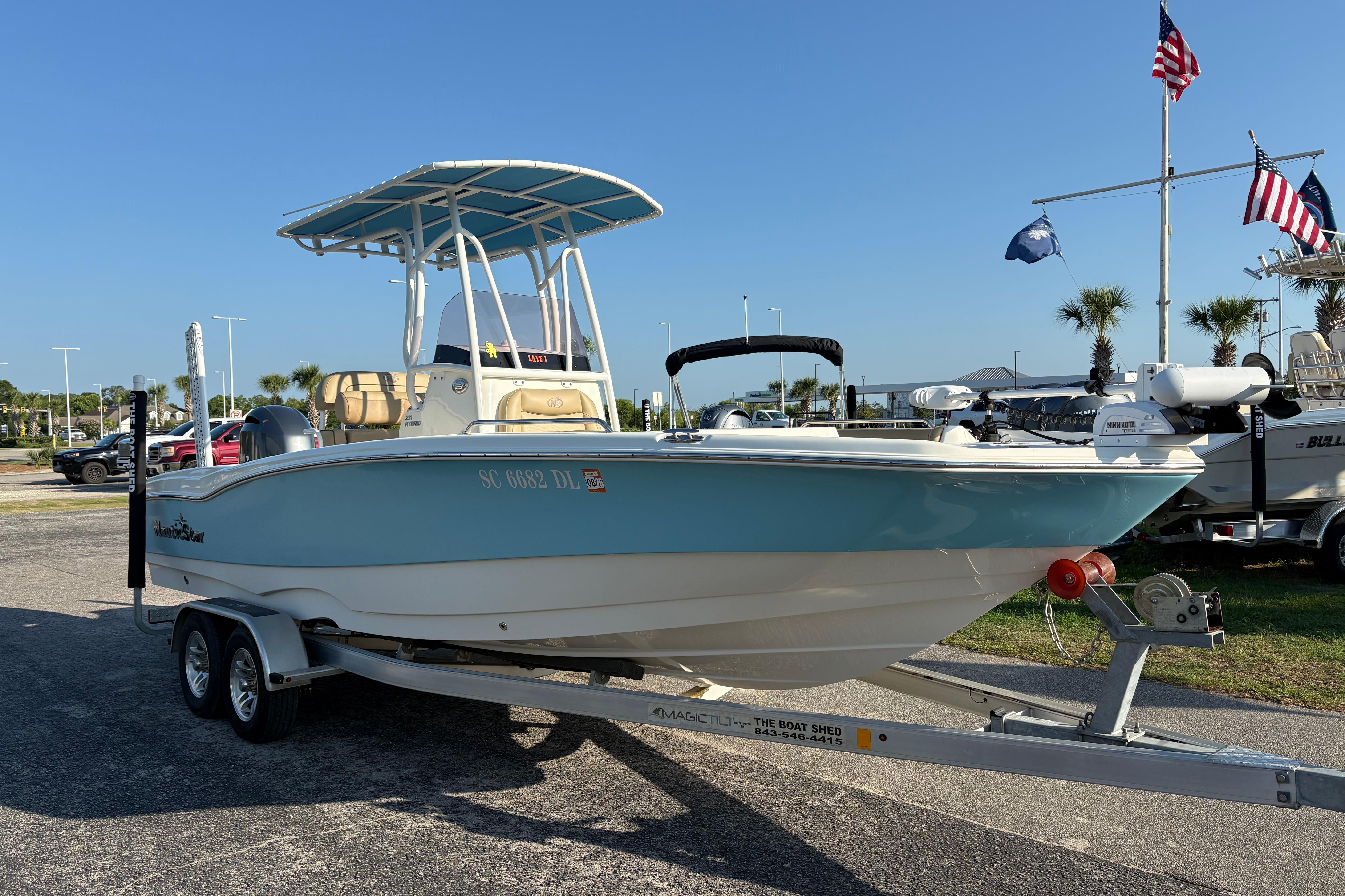 2019 NauticStar 231 Hybrid boat on trailer, displayed outdoors with flags in the background.