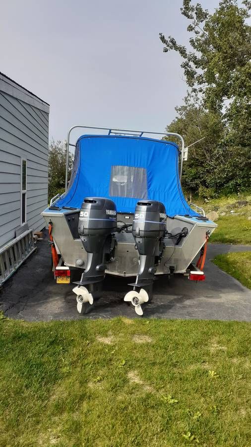 A boat with two outboard motors is on a trailer next to a house.