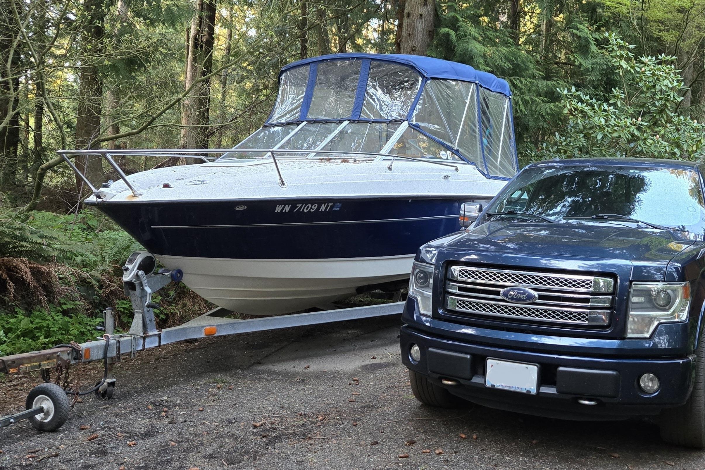 2006 Bayliner 192 Discovery Cuddy on trailer beside a parked truck in a wooded area.