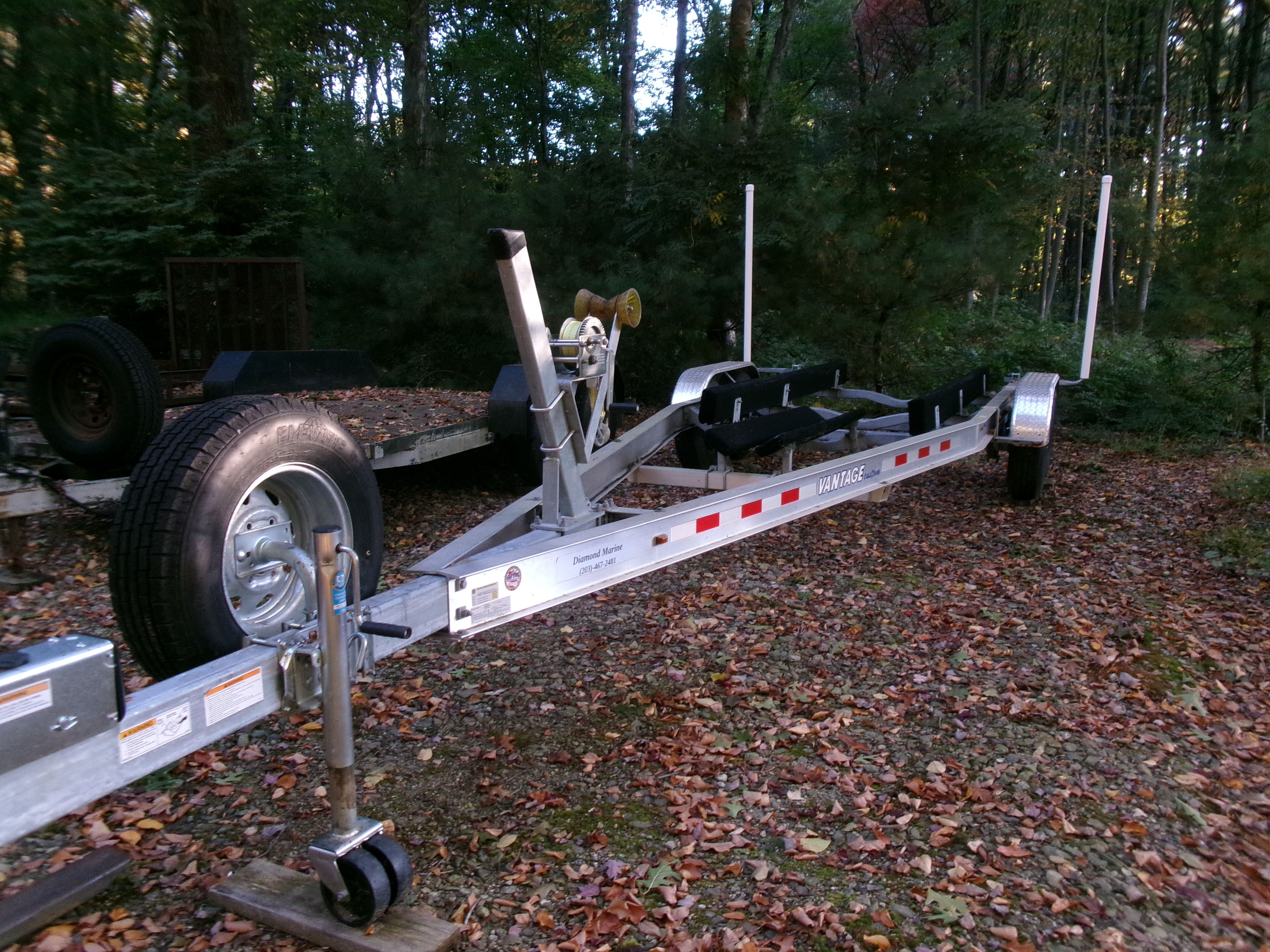 A boat trailer sits on a leaf-covered ground in a wooded area.