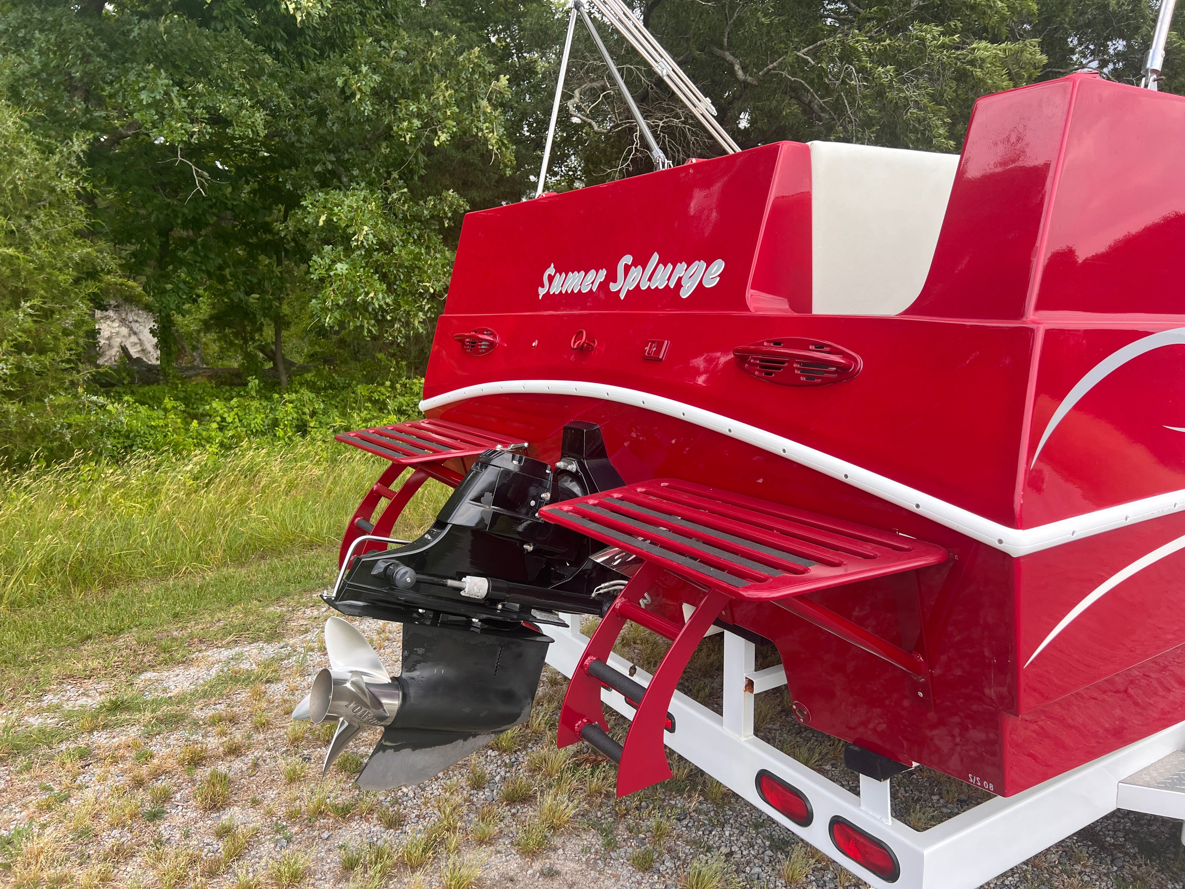 A red boat named "Summer Splurge" is on a trailer in a green setting.