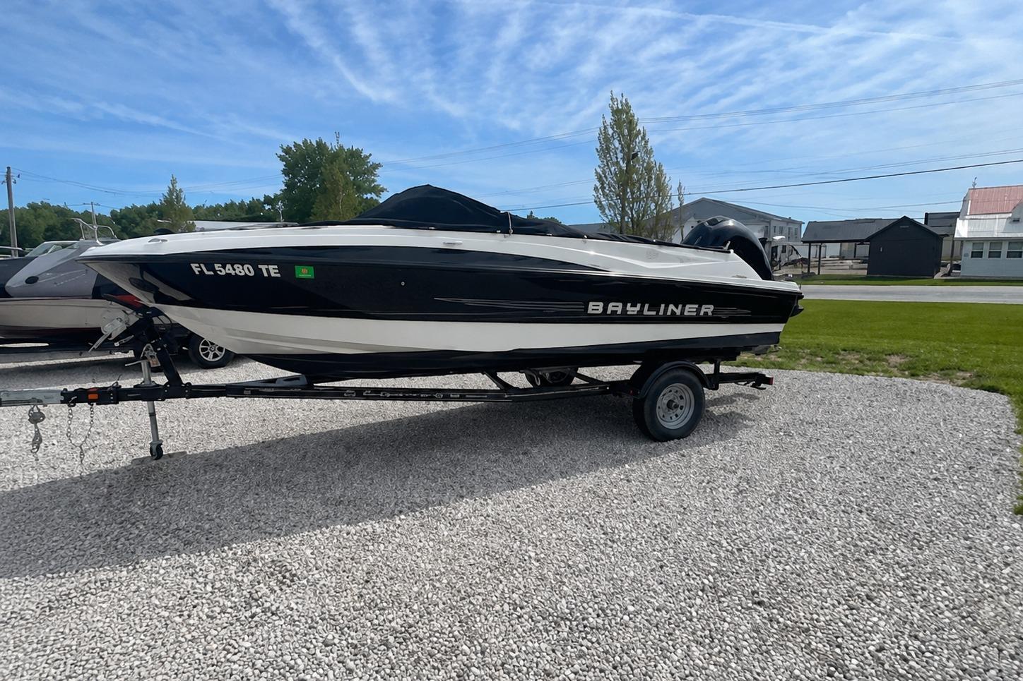 2013 Bayliner 190 Bowrider boat on trailer, parked on gravel, under clear blue sky.