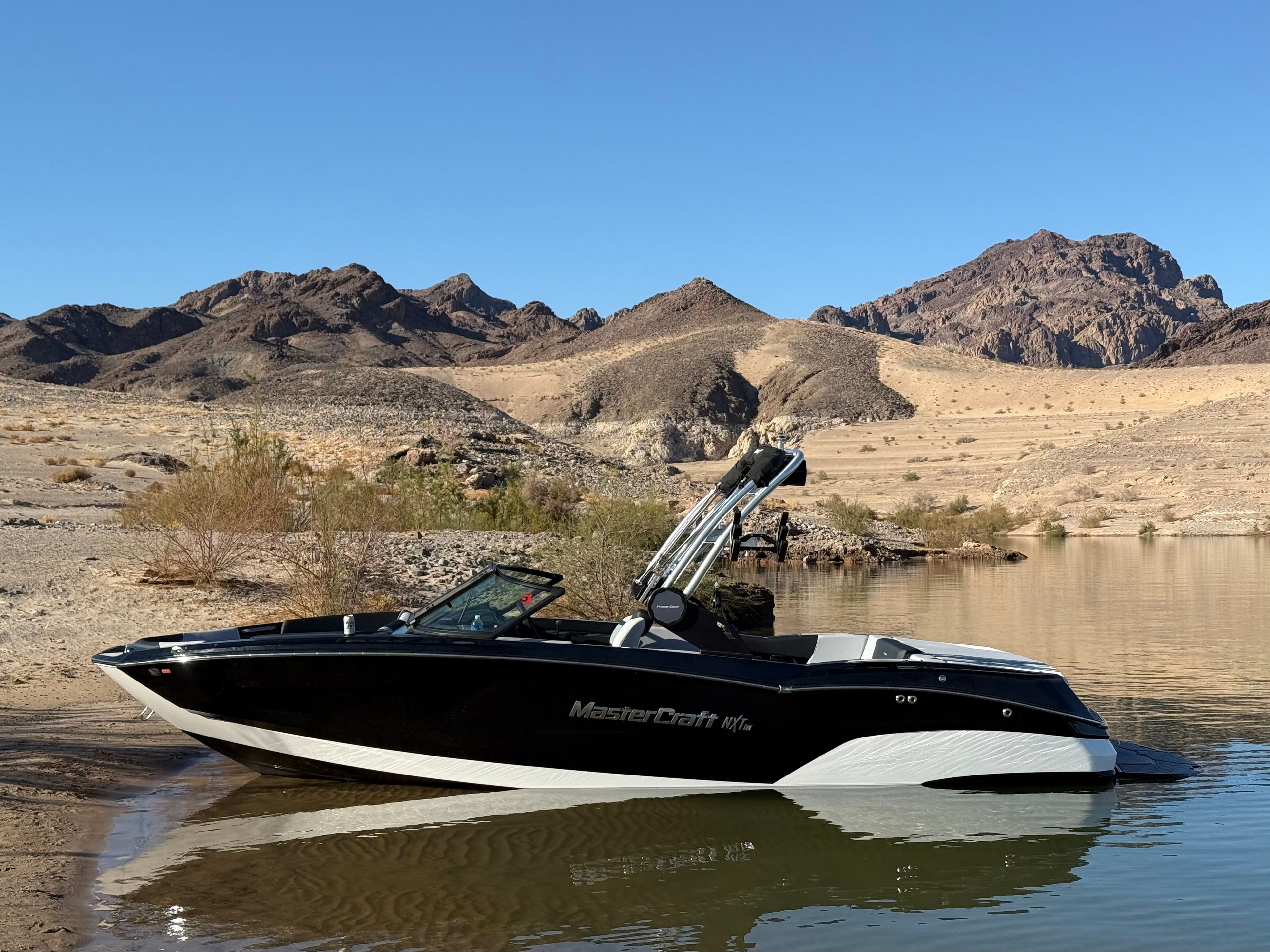 A MasterCraft NXT23 boat is docked by a calm lake in a desert landscape.