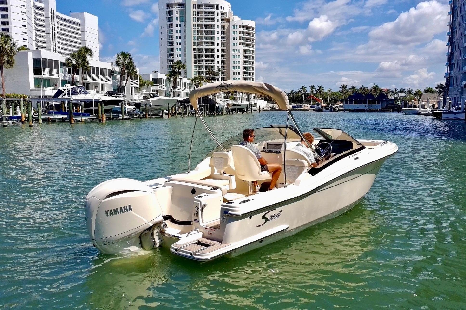 2026 Scout 220 Dorado boat cruising in a sunny marina with modern buildings.