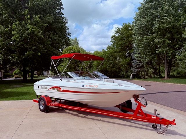 A white boat with red accents on a trailer, surrounded by trees and a blue sky.
