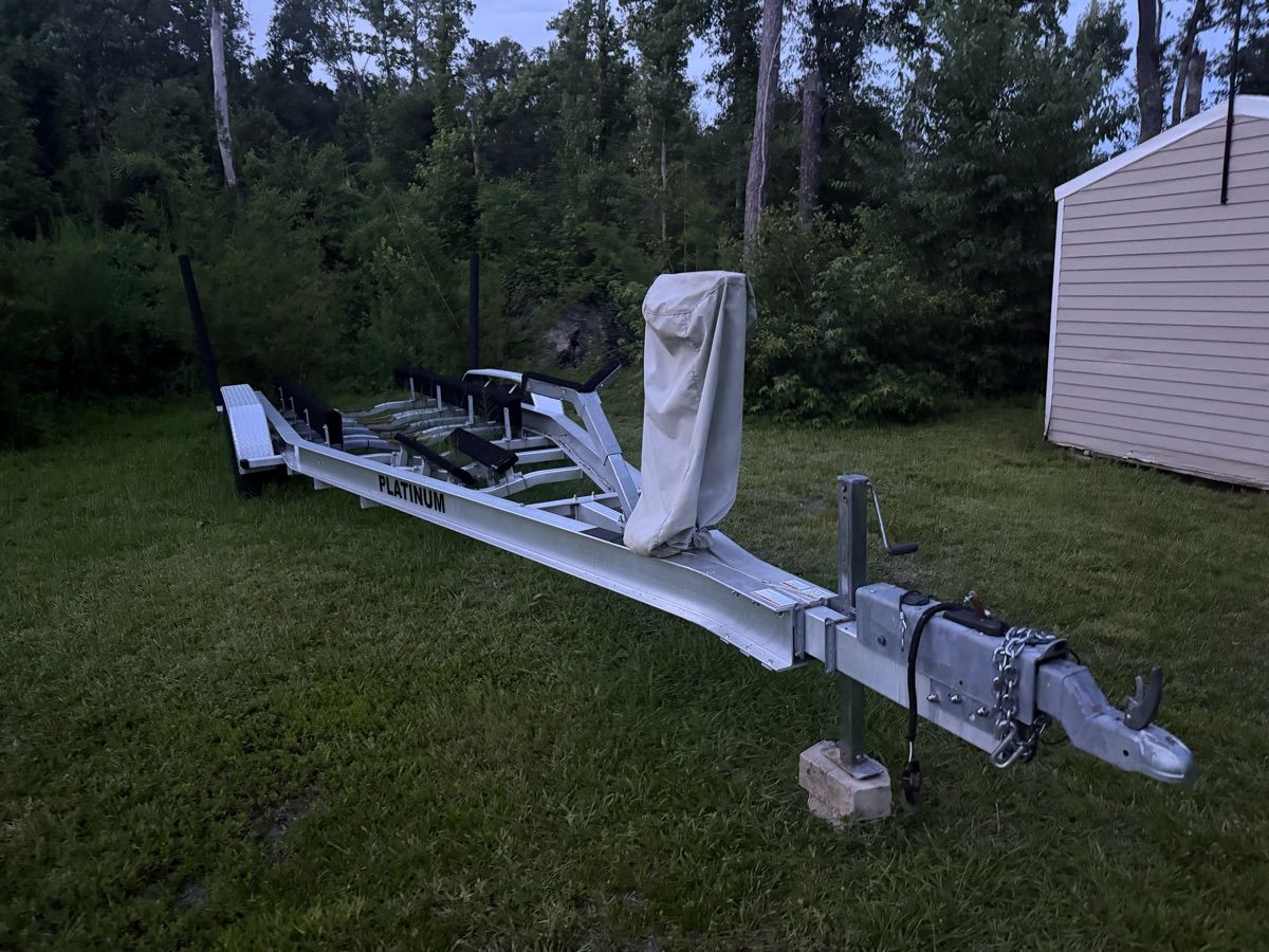 A boat trailer sits on grass, surrounded by trees.