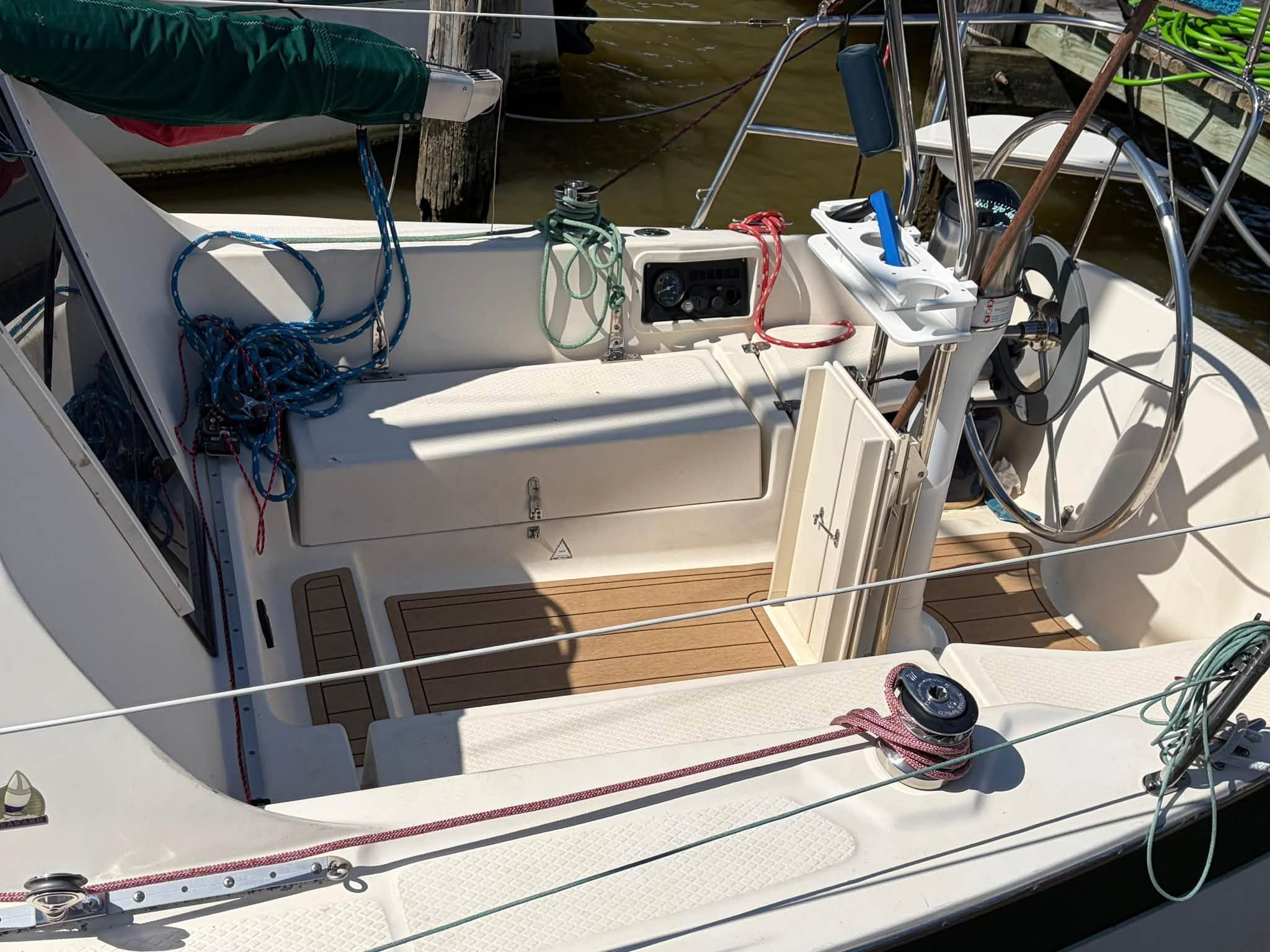 The cockpit of a sailboat with a steering wheel, ropes, and wooden flooring.