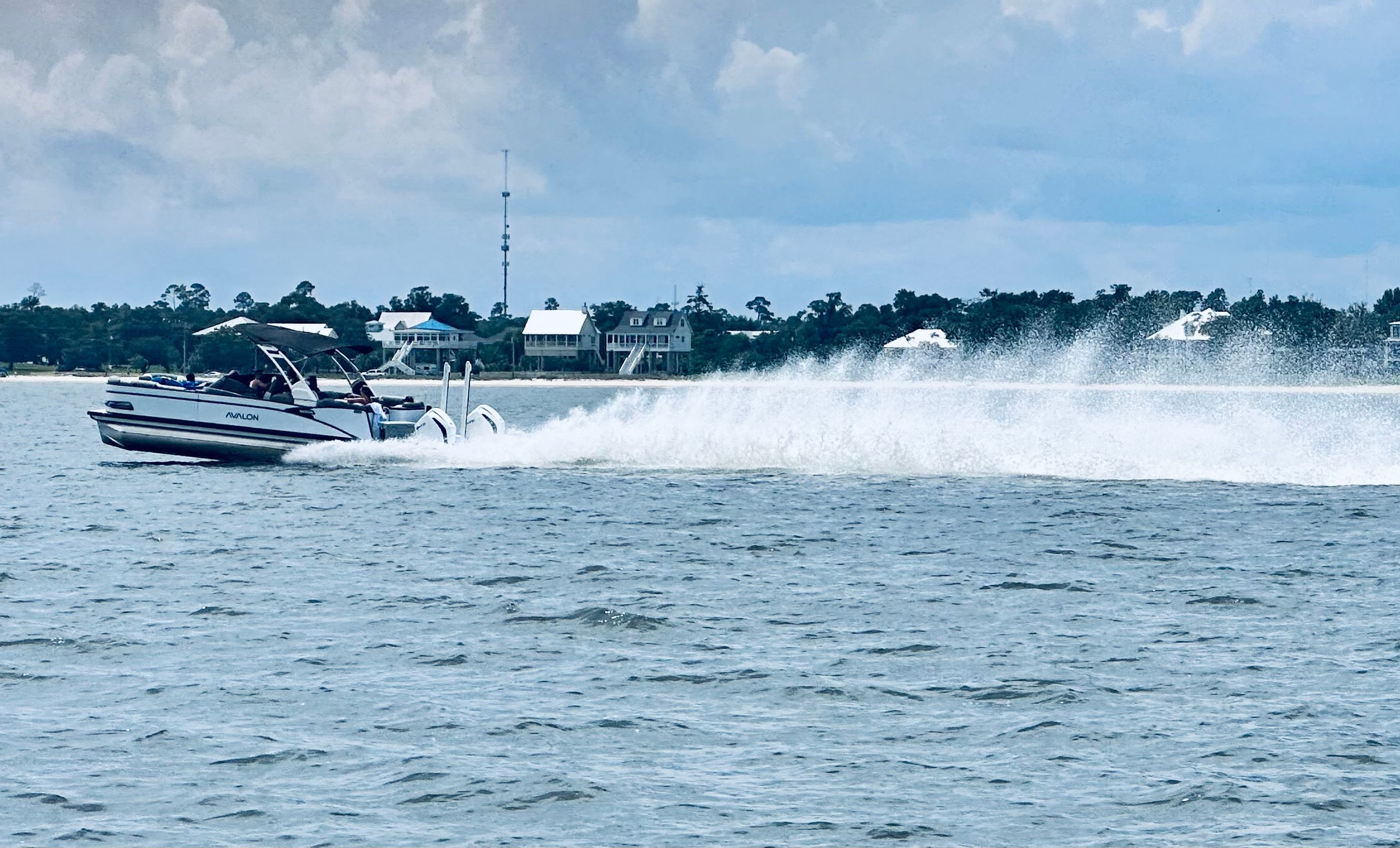 A 2024 Avalon Catalina Quad Lounger speeds across the water, with houses visible in the background.