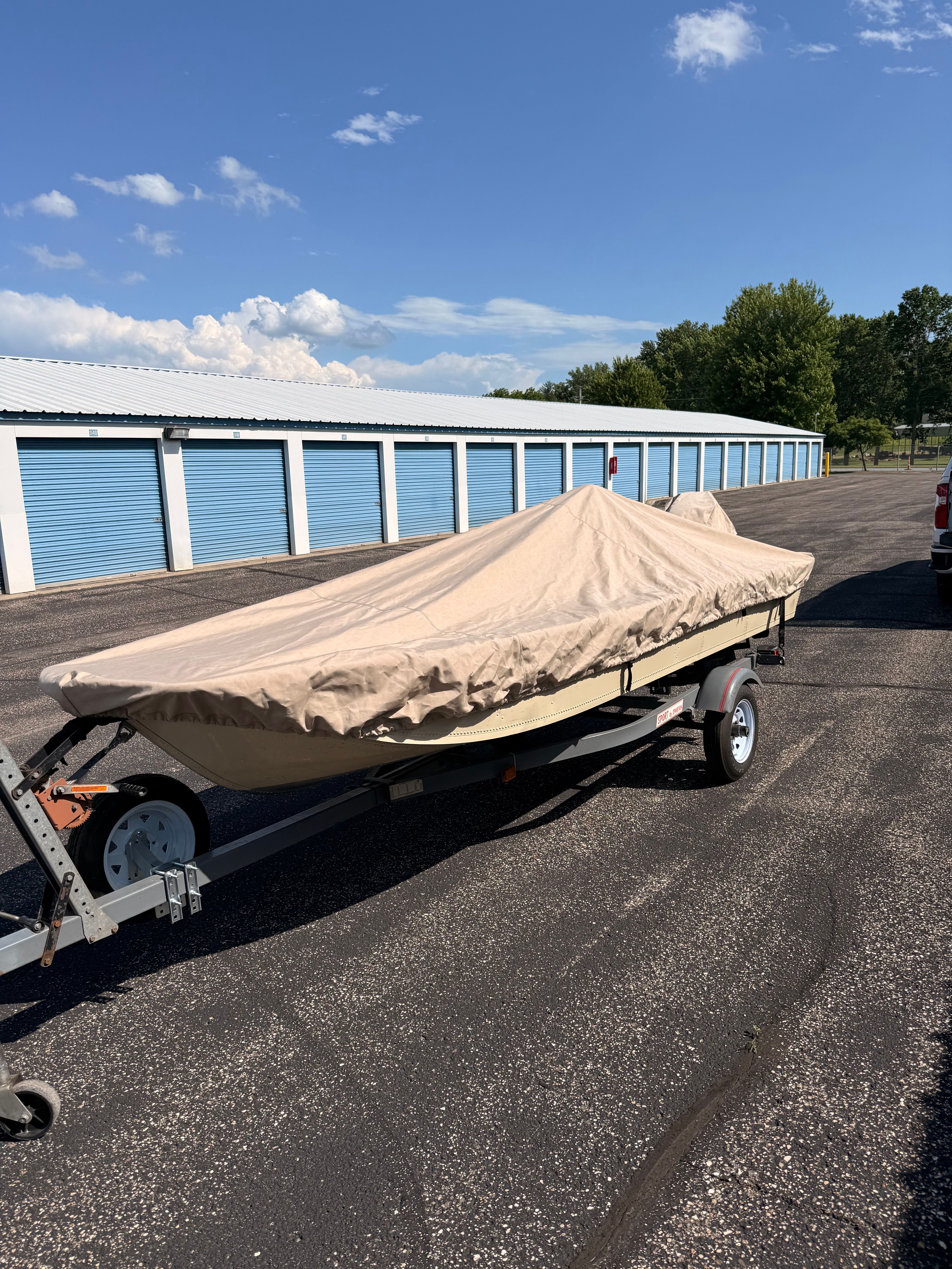 A 1985 Grumman 4.4 boat on a trailer, covered with a tarp, parked on a lot.