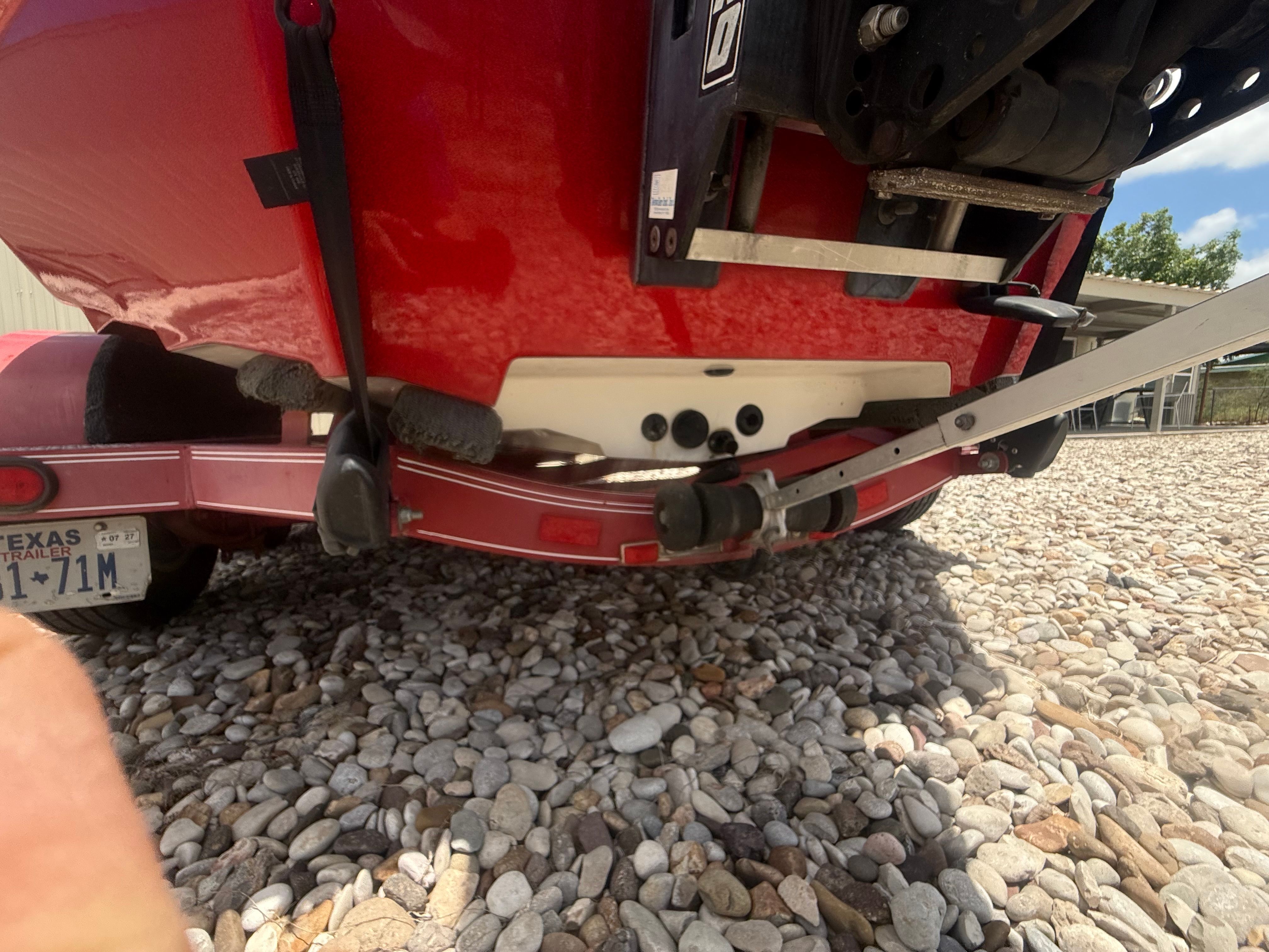 The image shows the rear of a red boat on a trailer, surrounded by pebbles.