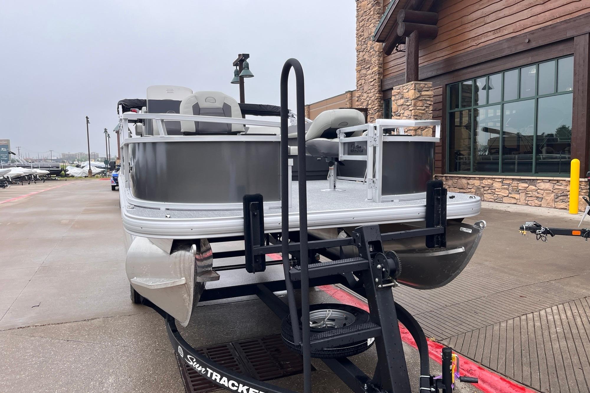 2026 Sun Tracker Bass Buggy 16 XL Select pontoon boat on trailer near rustic building.
