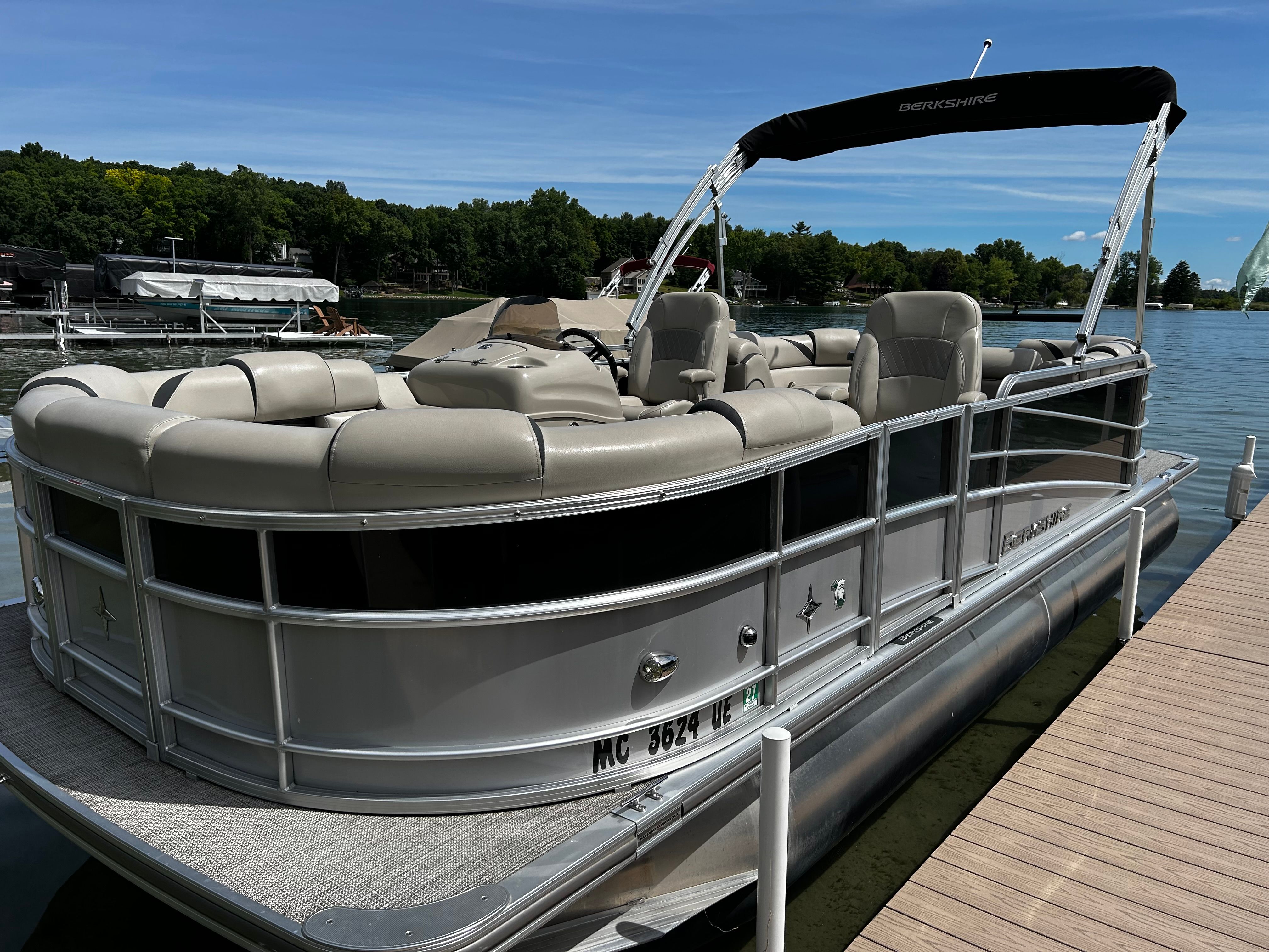A sleek pontoon boat docked at a wooden pier, surrounded by calm water and green trees.