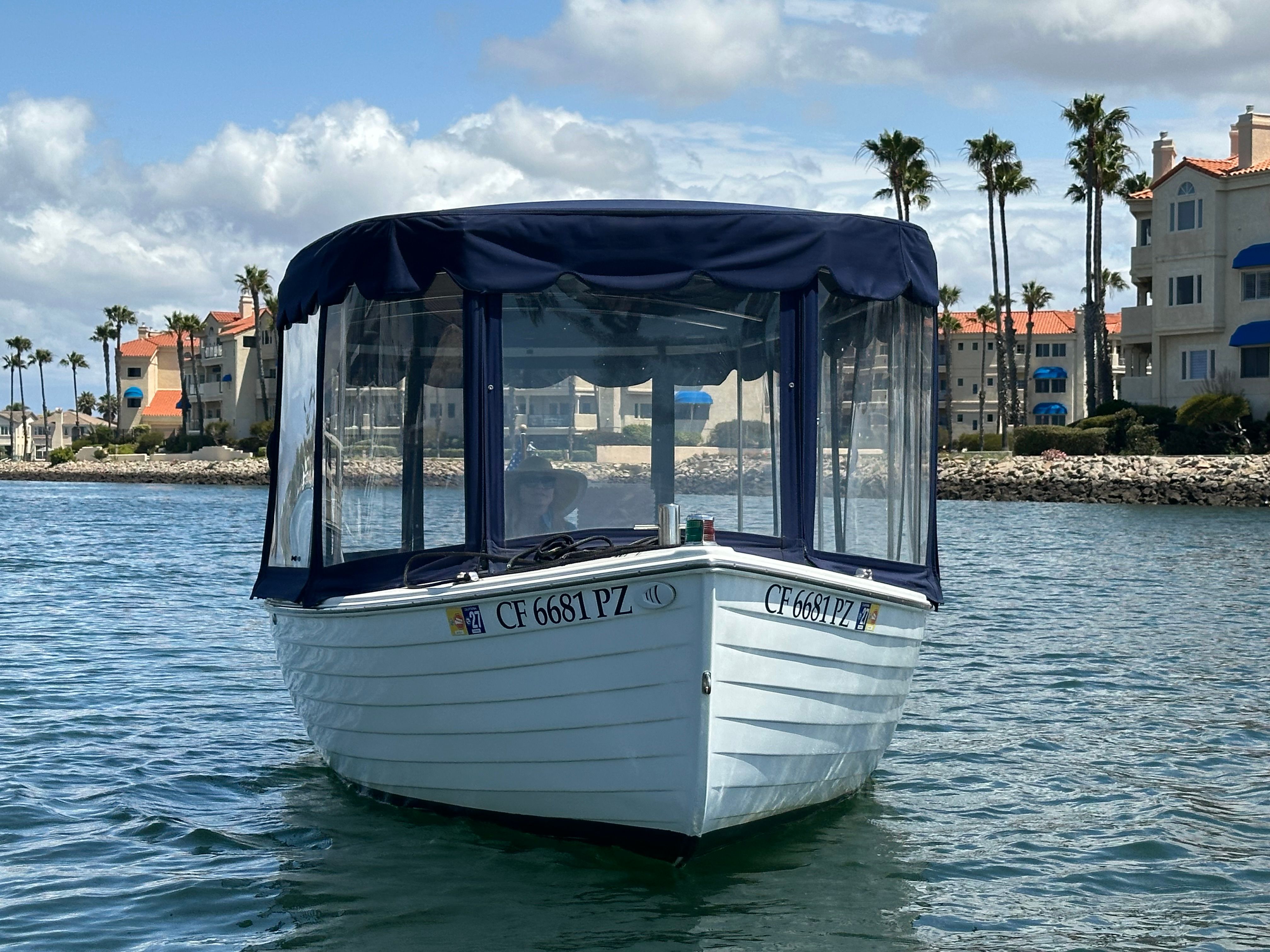 A white boat with a blue canopy on calm water, surrounded by palm trees and buildings.