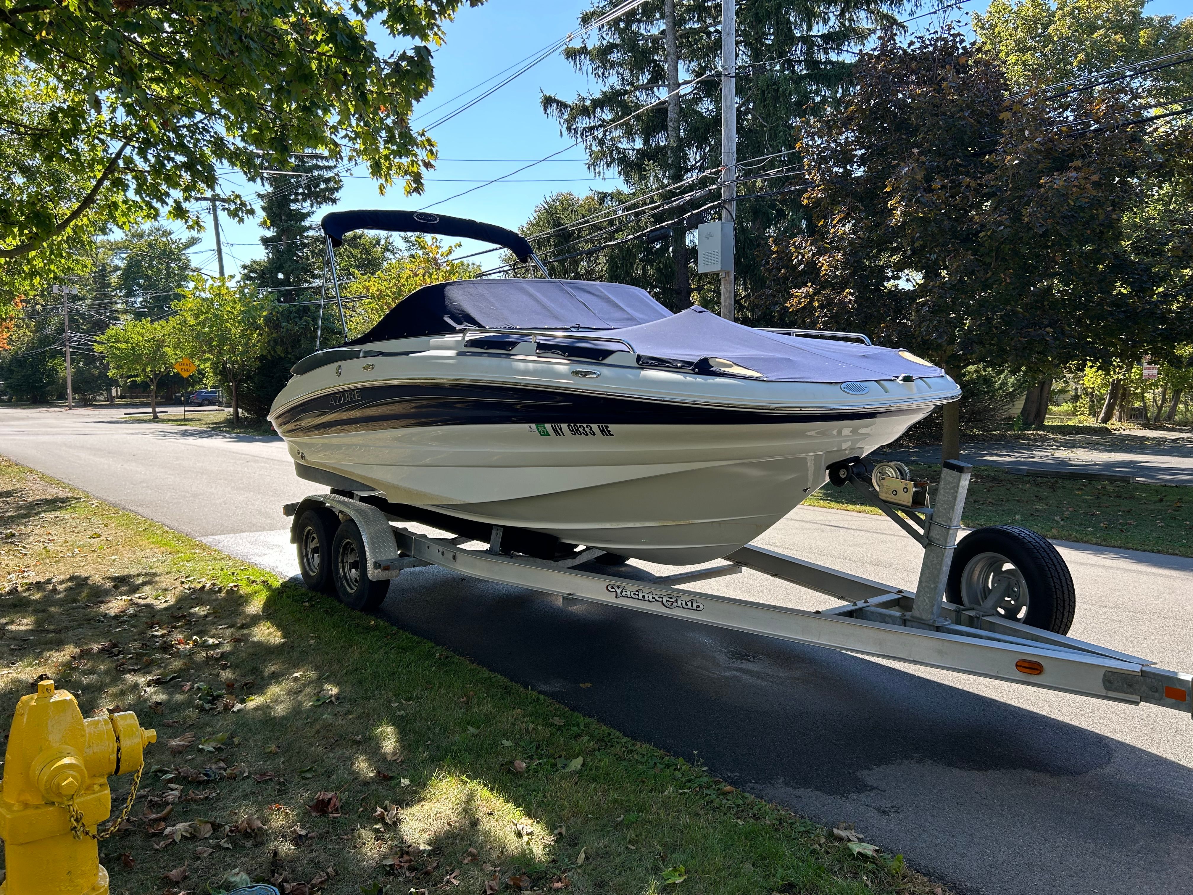 A white boat parked on a trailer on a street, surrounded by trees.