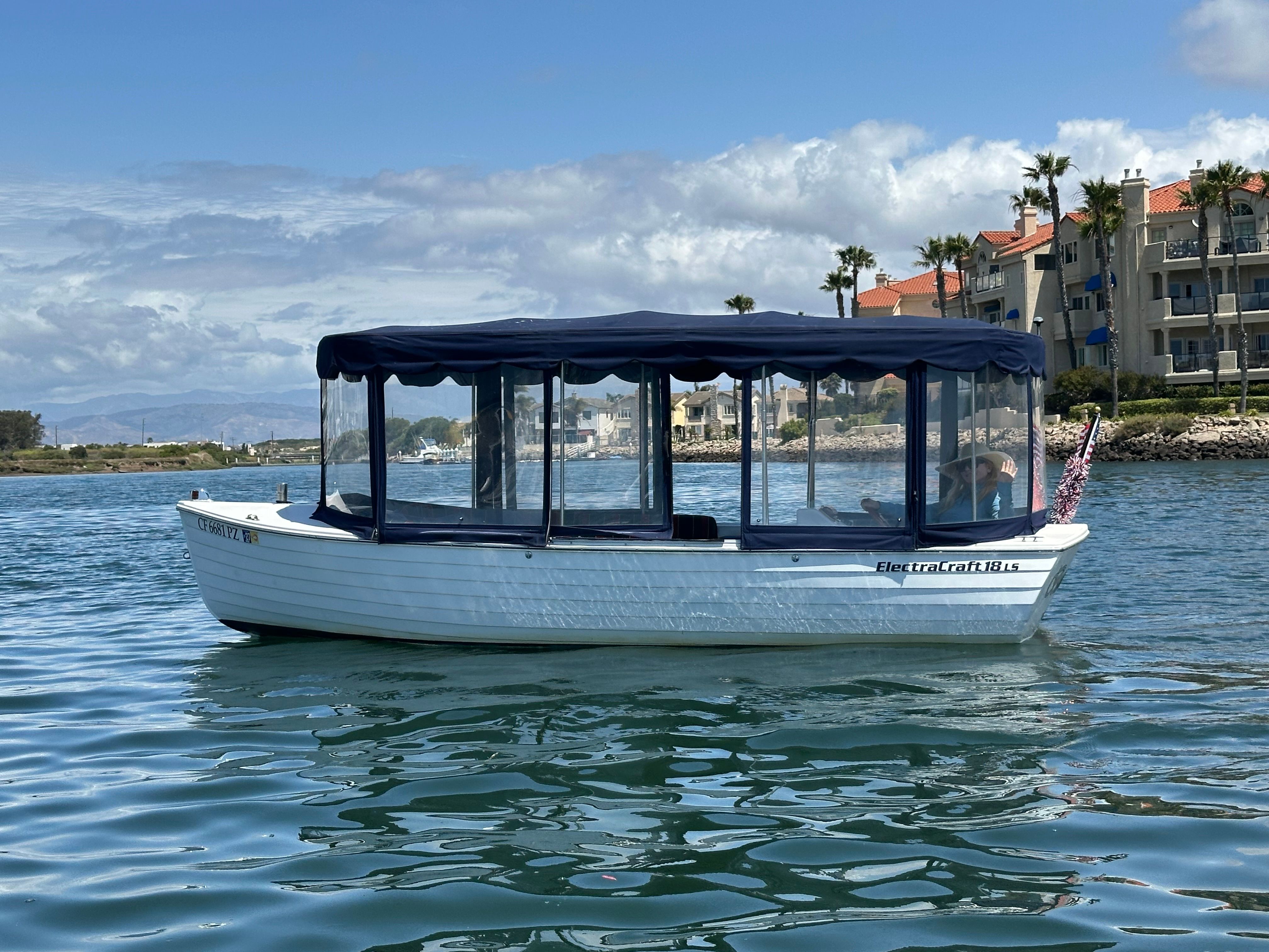 A white boat with a blue canopy on calm water near a shoreline with buildings.
